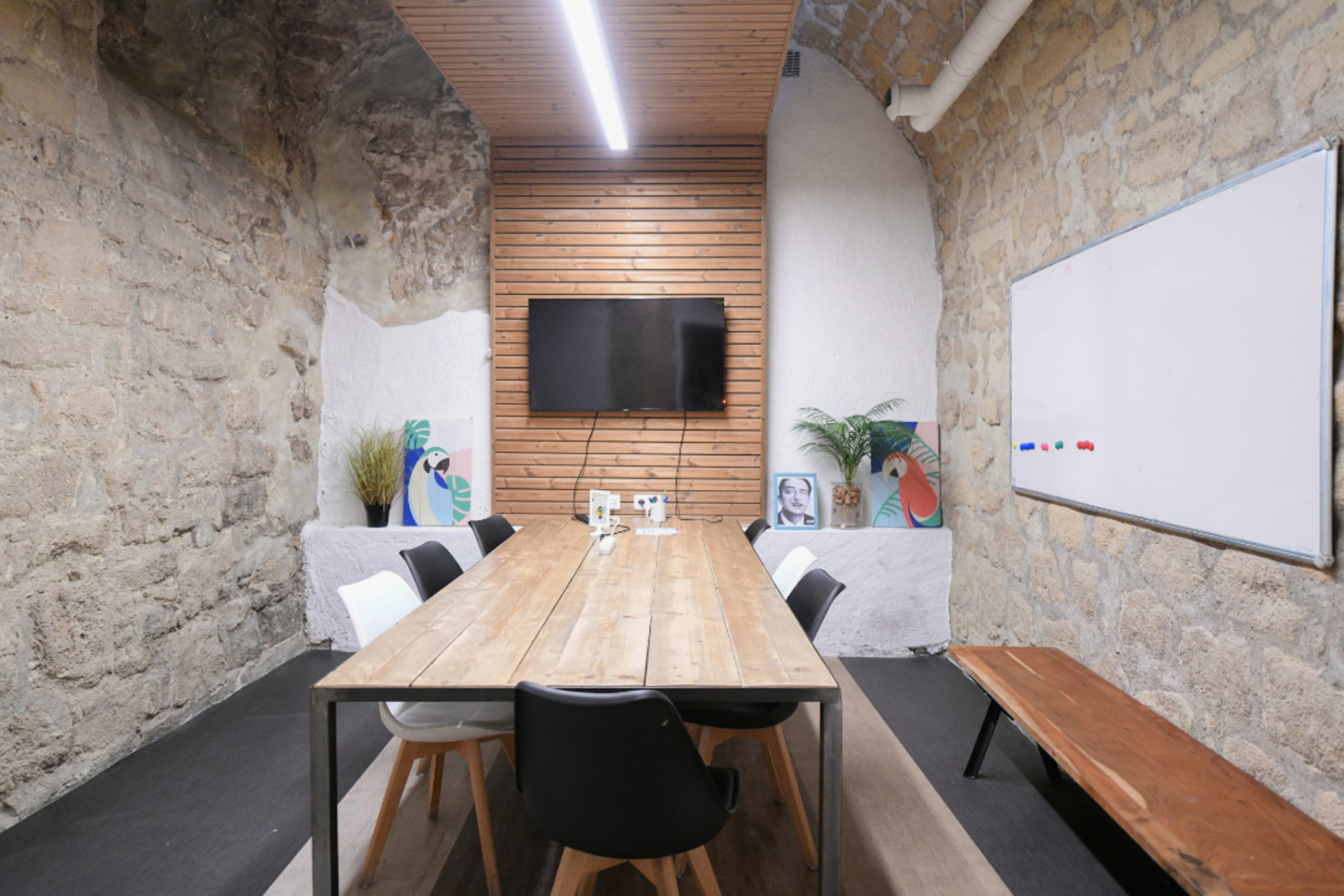 A modern conference room features a long wooden table, black and white chairs, a television mounted on a wooden wall, and artworks displayed on the stone walls.