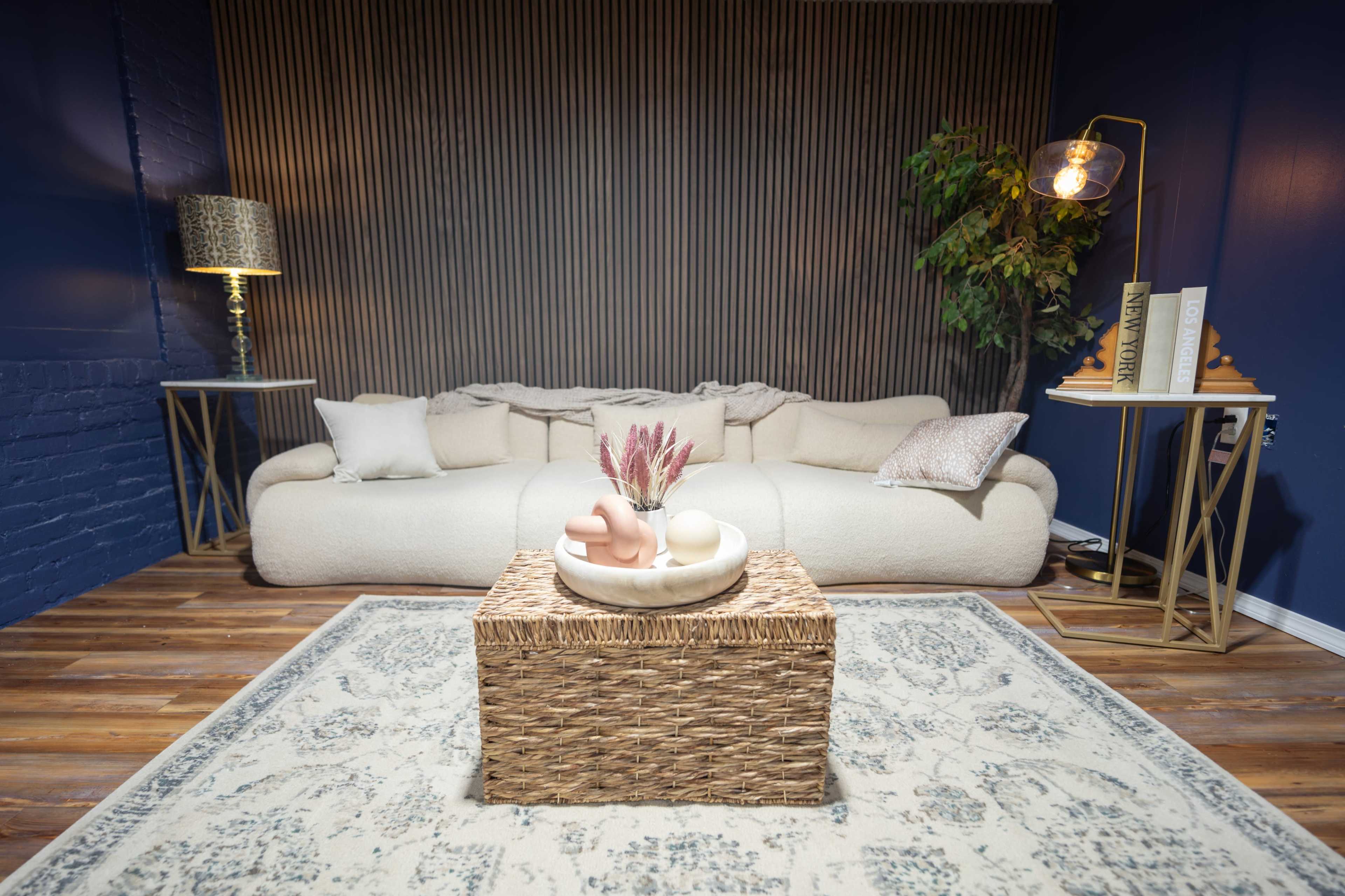 The image shows a cozy living room featuring a curved, light-colored sofa, a woven coffee table, and decor elements including a plant and a table lamp.