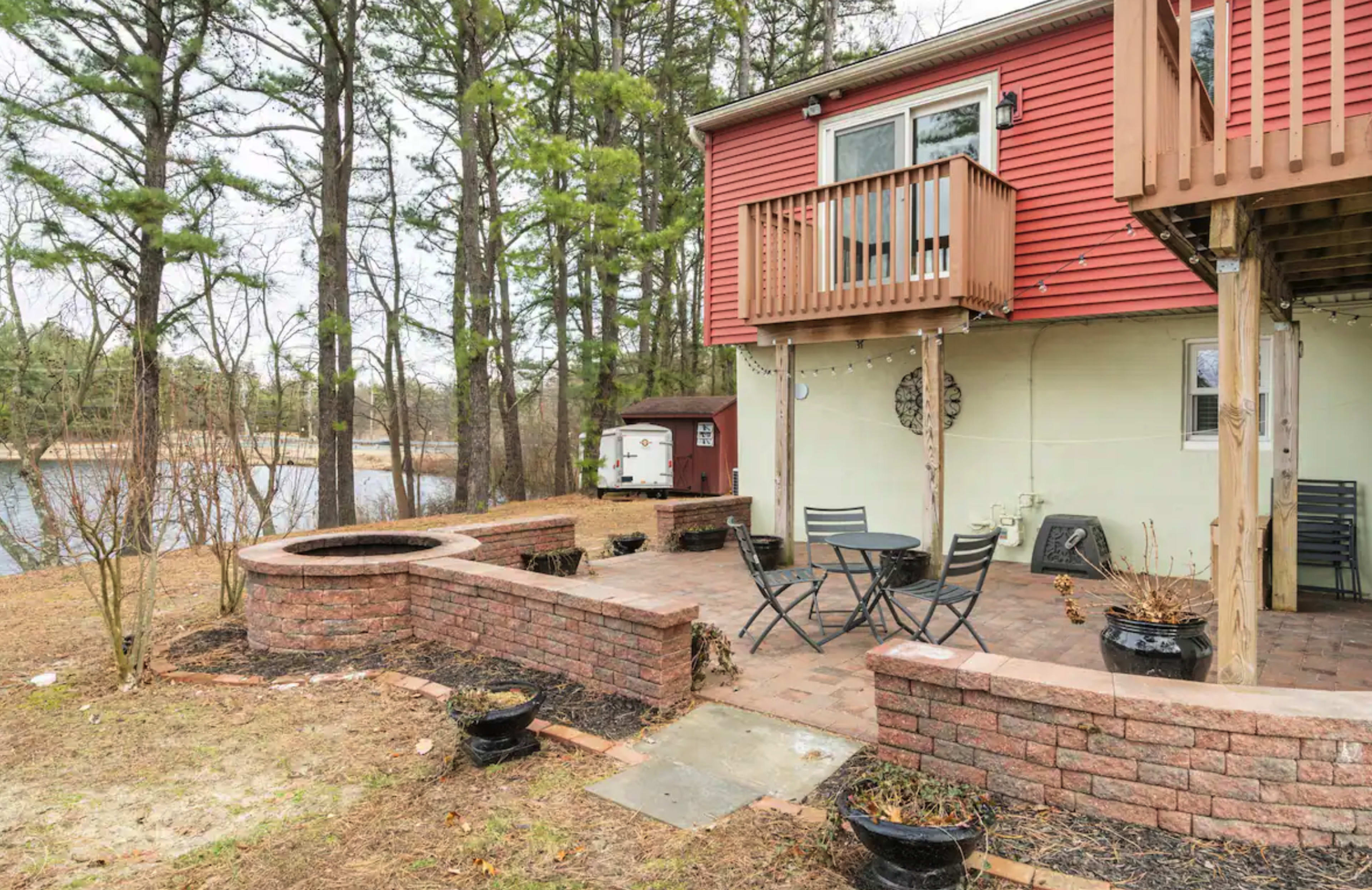 A brick patio with black chairs and a circular fire pit is situated next to a house by a body of water, surrounded by trees.