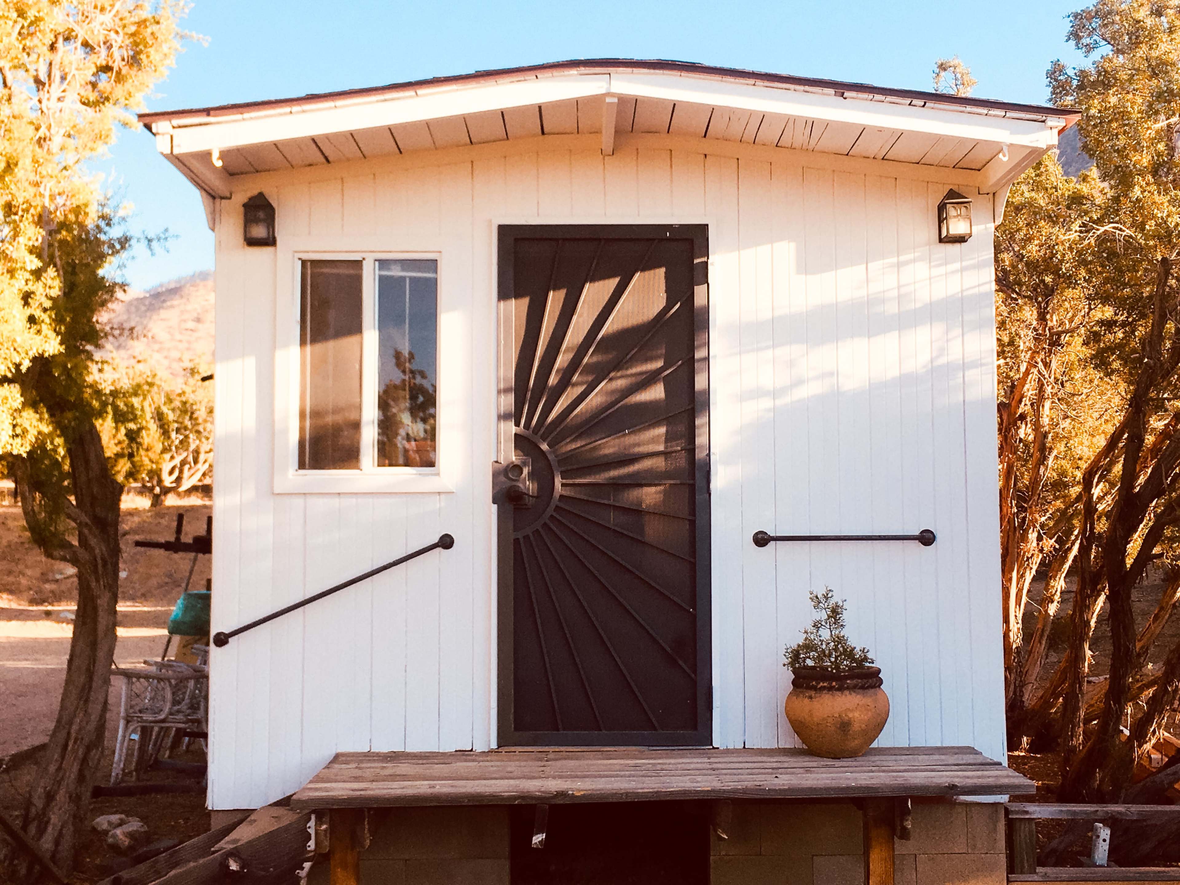 A small white cabin with a distinctive spiral door design, situated on a wooden platform amid trees.