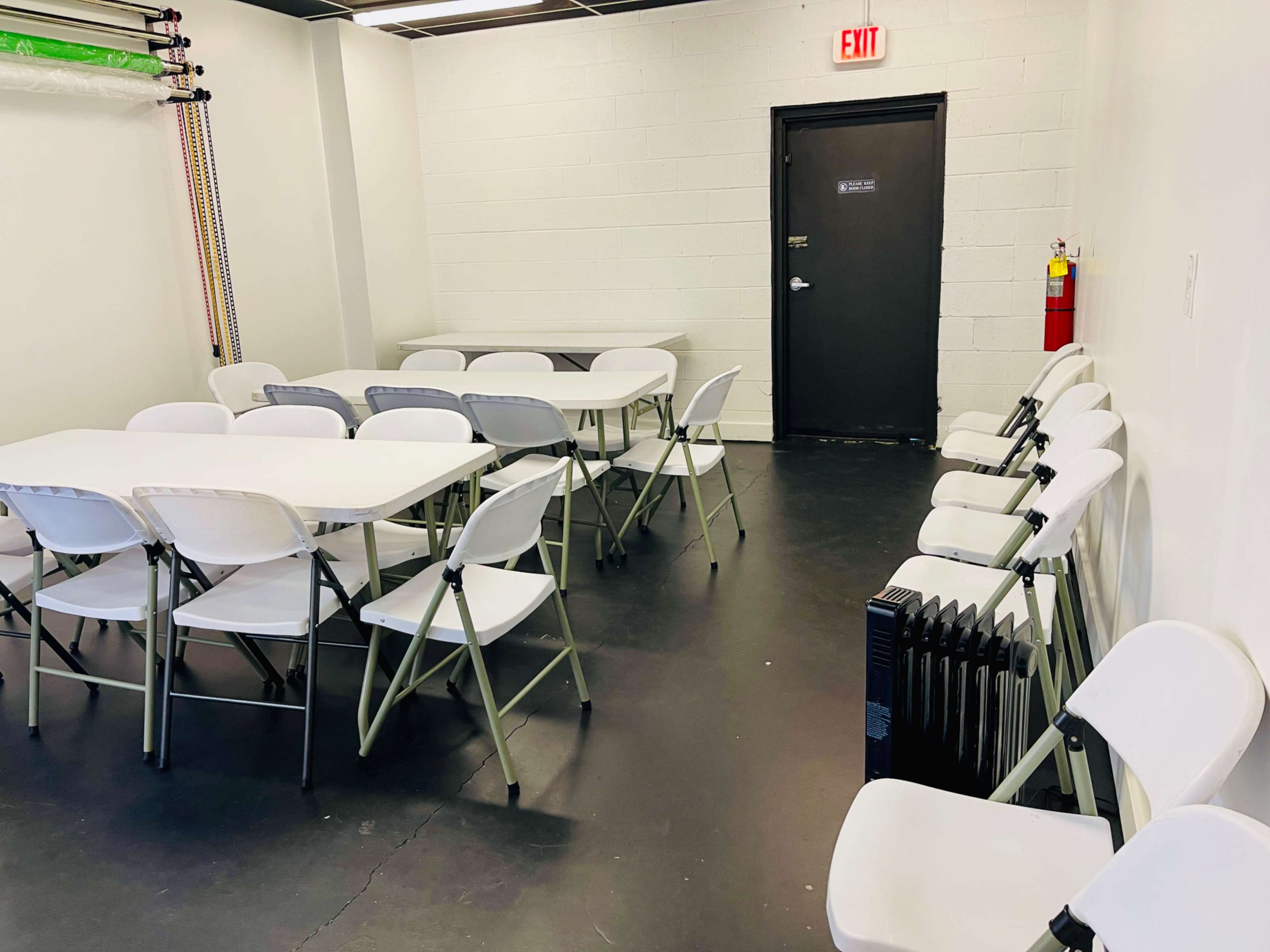 A room with several white folding tables and chairs arranged around, a black door, and a radiator along the wall.