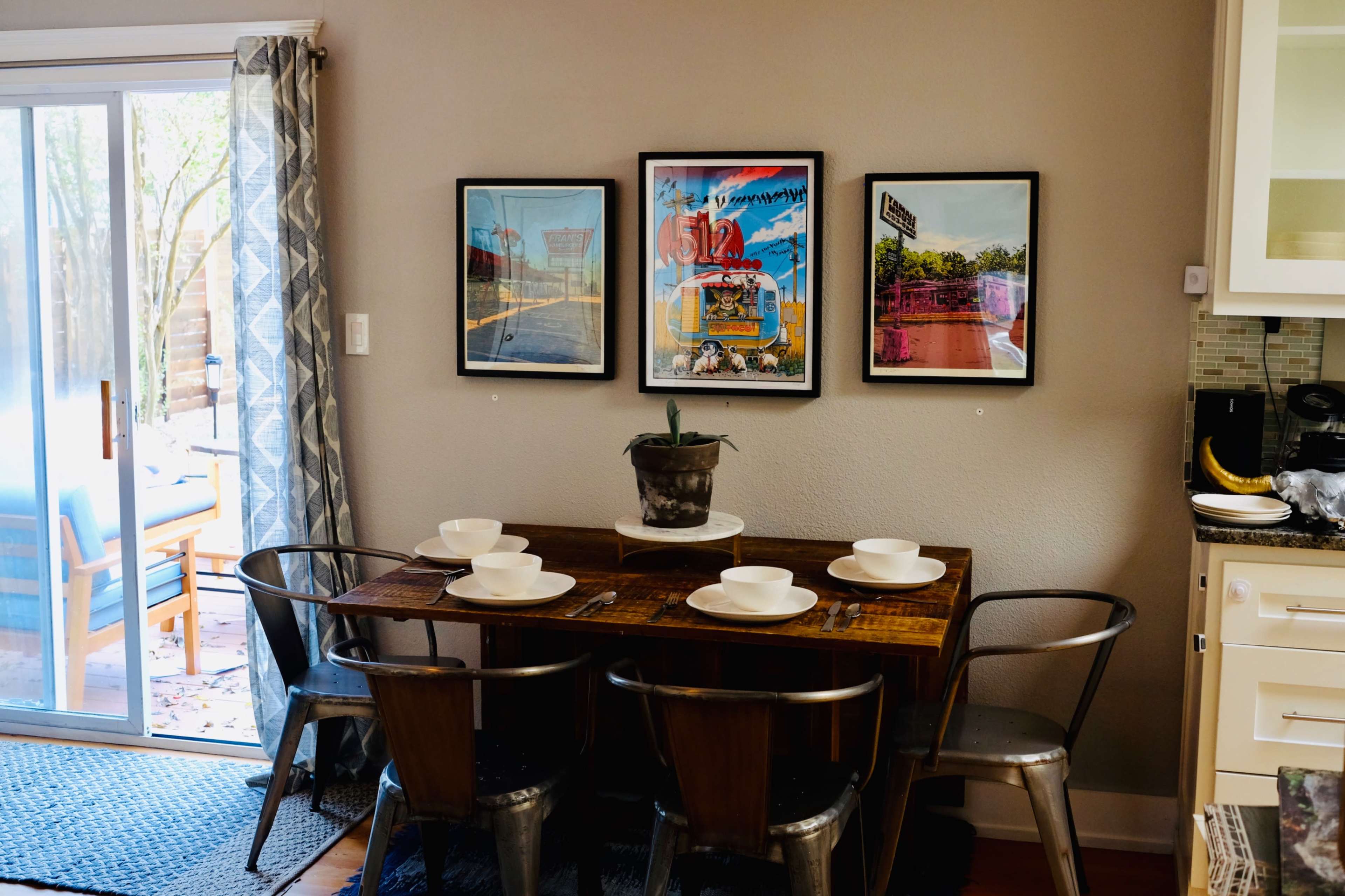 A wooden dining table with four white bowls sits in a well-lit kitchen area, complemented by framed artwork on the wall and an outdoor view through a sliding glass door.