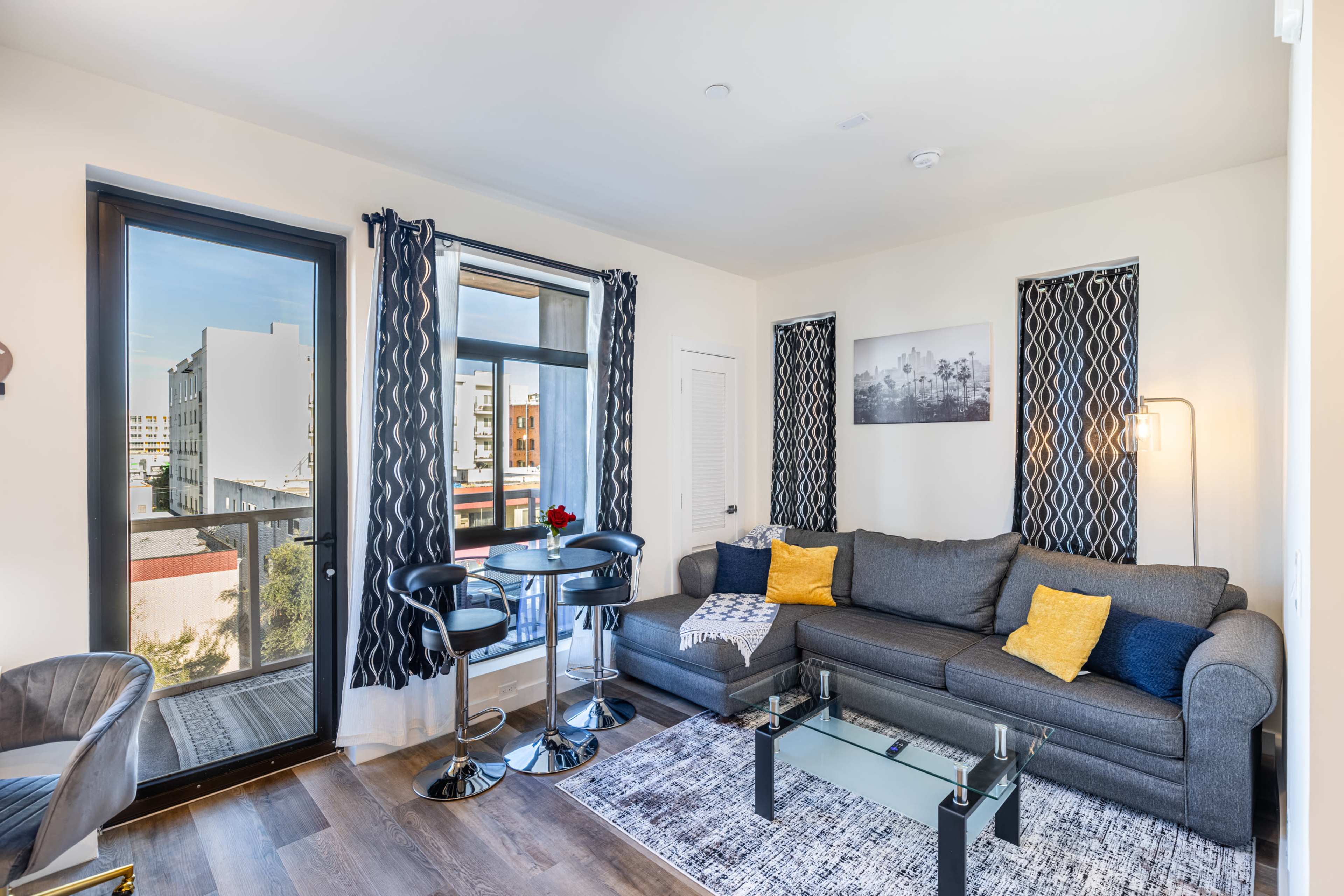 A cozy living room features a gray couch, a glass coffee table, and a small dining area with two chairs, complemented by large windows and modern decor.