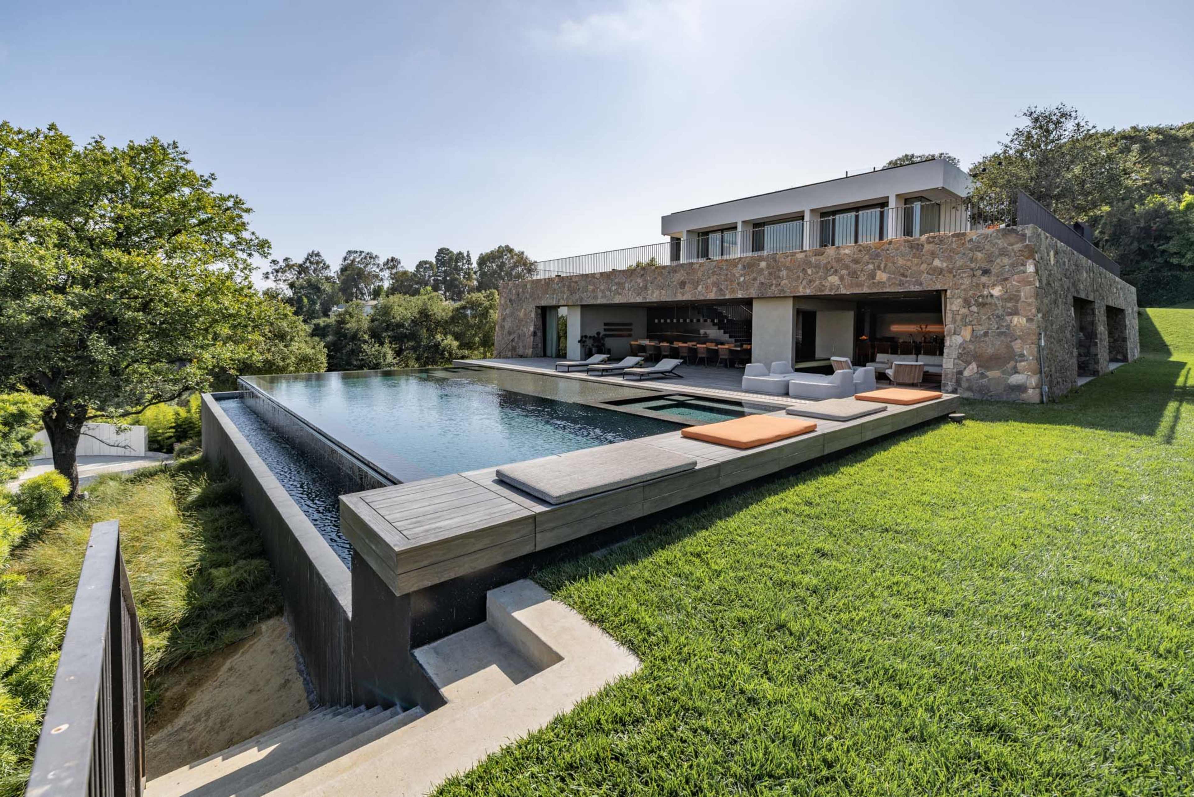 The image shows a modern house with large windows overlooking an infinity pool and a well-manicured lawn.