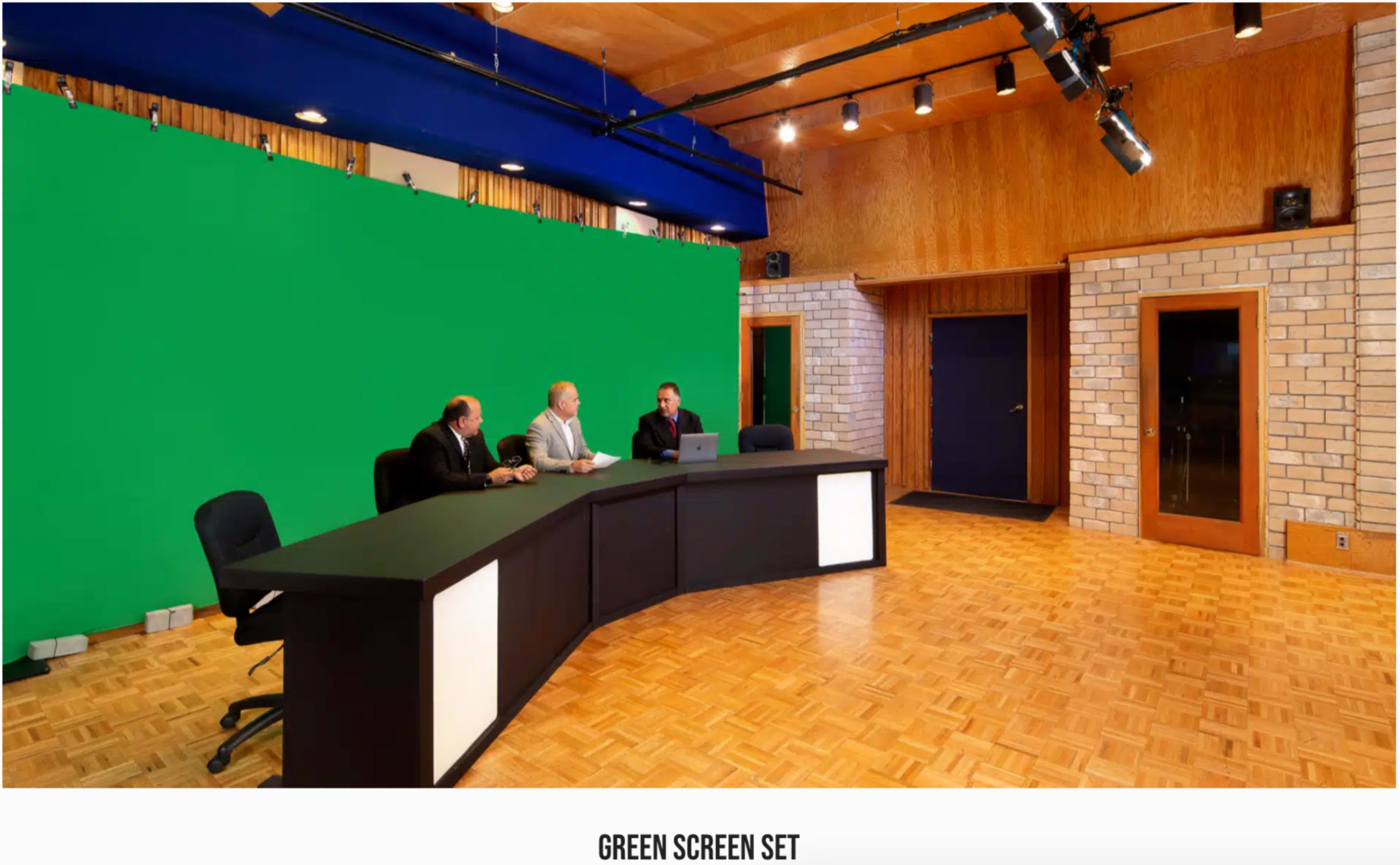 The image shows a green screen set with three men seated behind a black curved desk in a spacious room with wooden paneling and a patterned floor.