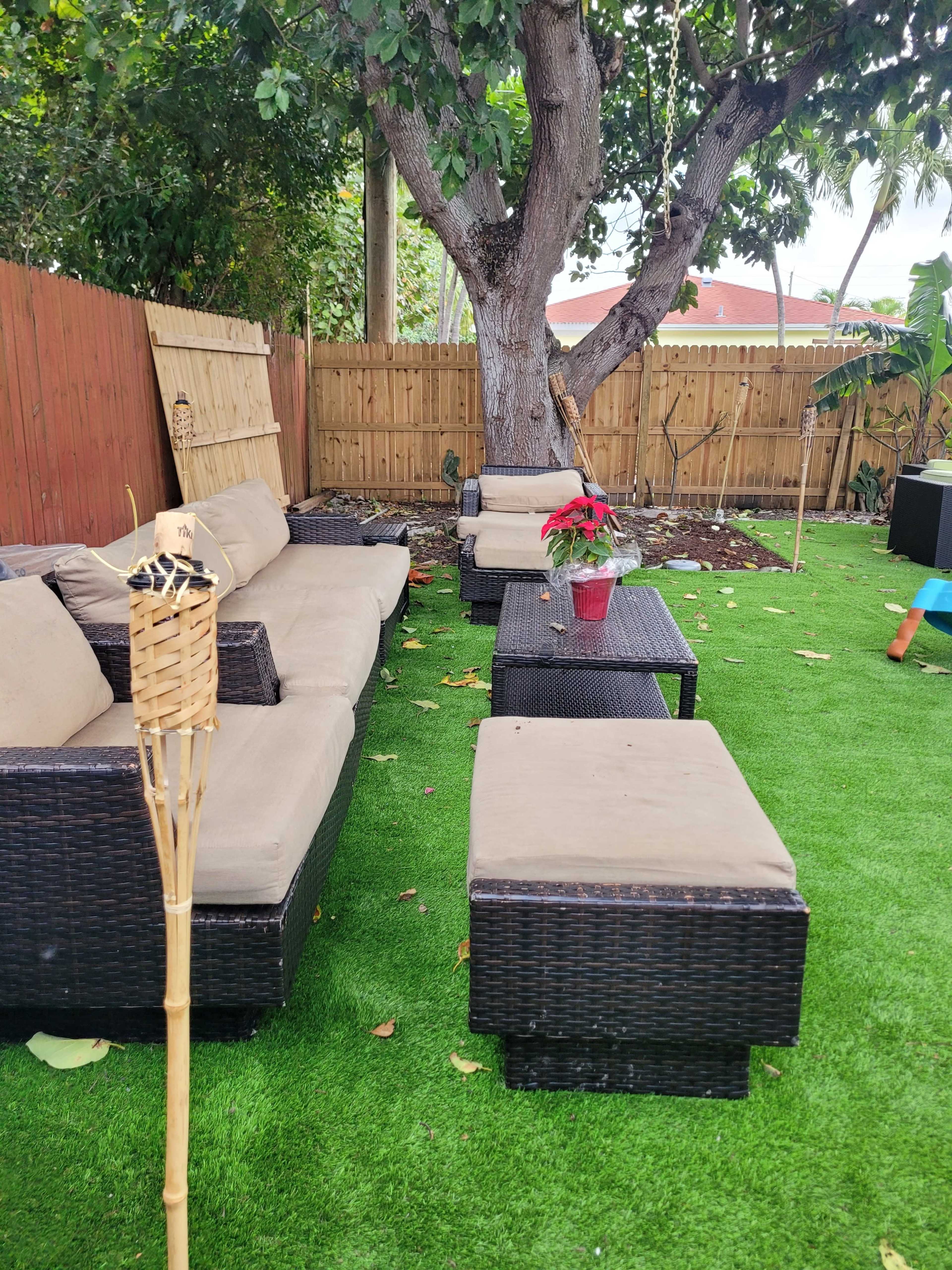 A backyard seating area features dark wicker furniture with beige cushions, surrounded by artificial grass and a wooden fence.
