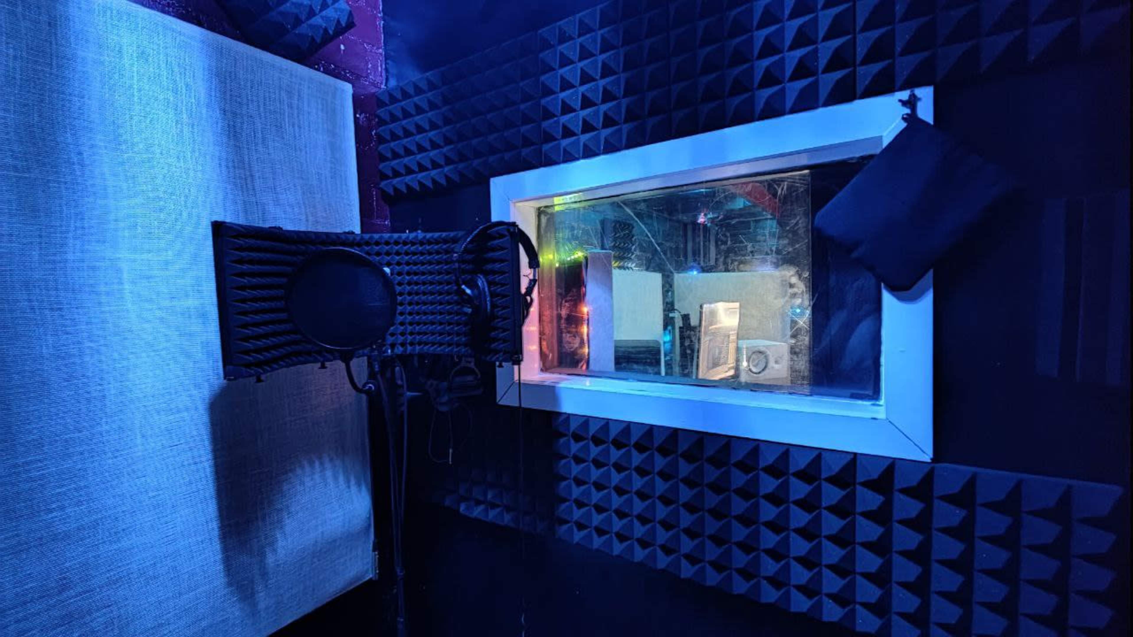 The image shows a soundproof recording booth with foam panels on the walls, a microphone stand, and a viewing window into a separate room.