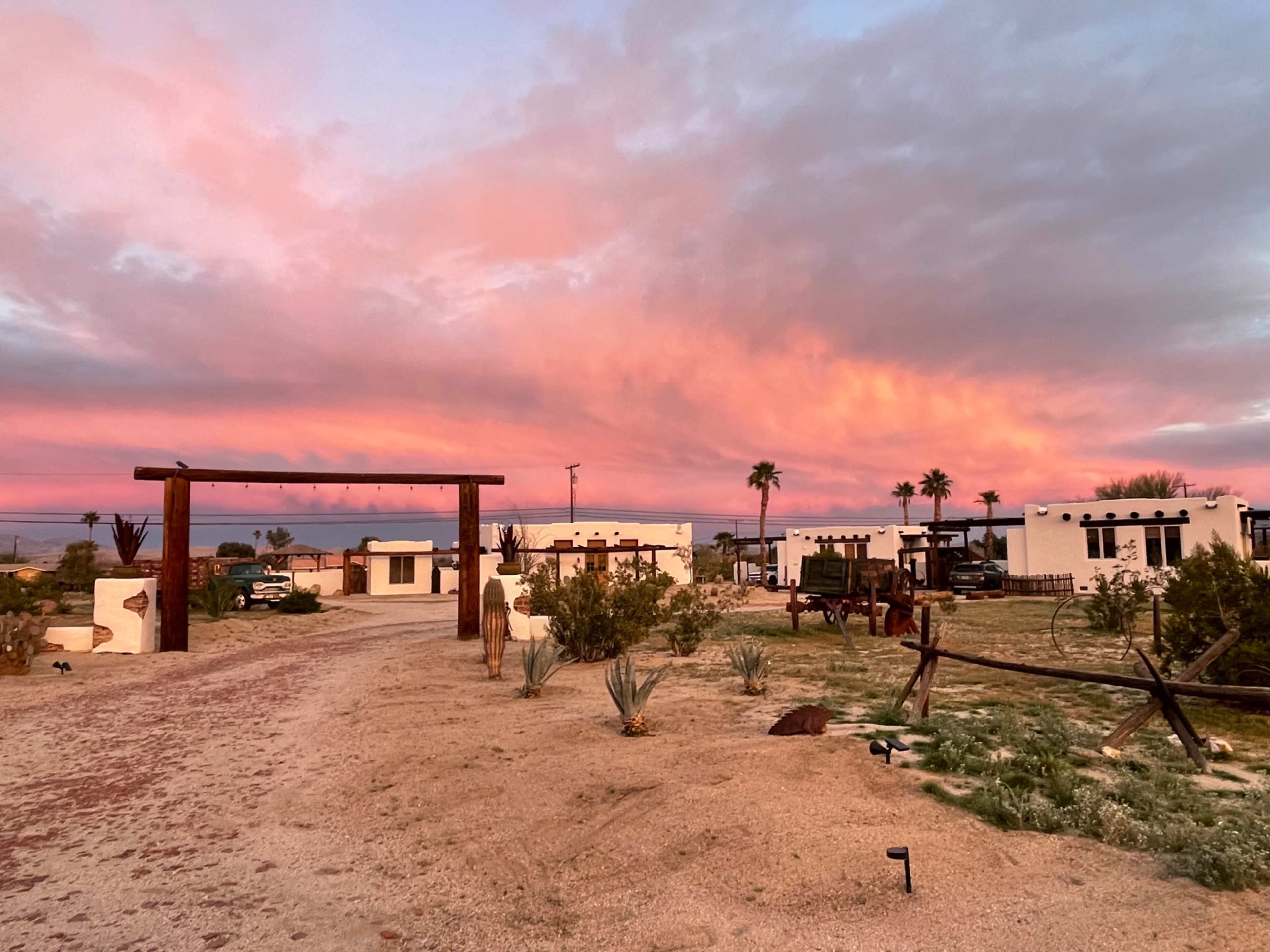 The scene depicts a desert landscape at sunset, featuring colorful clouds and several low white buildings surrounded by palm trees and desert vegetation.