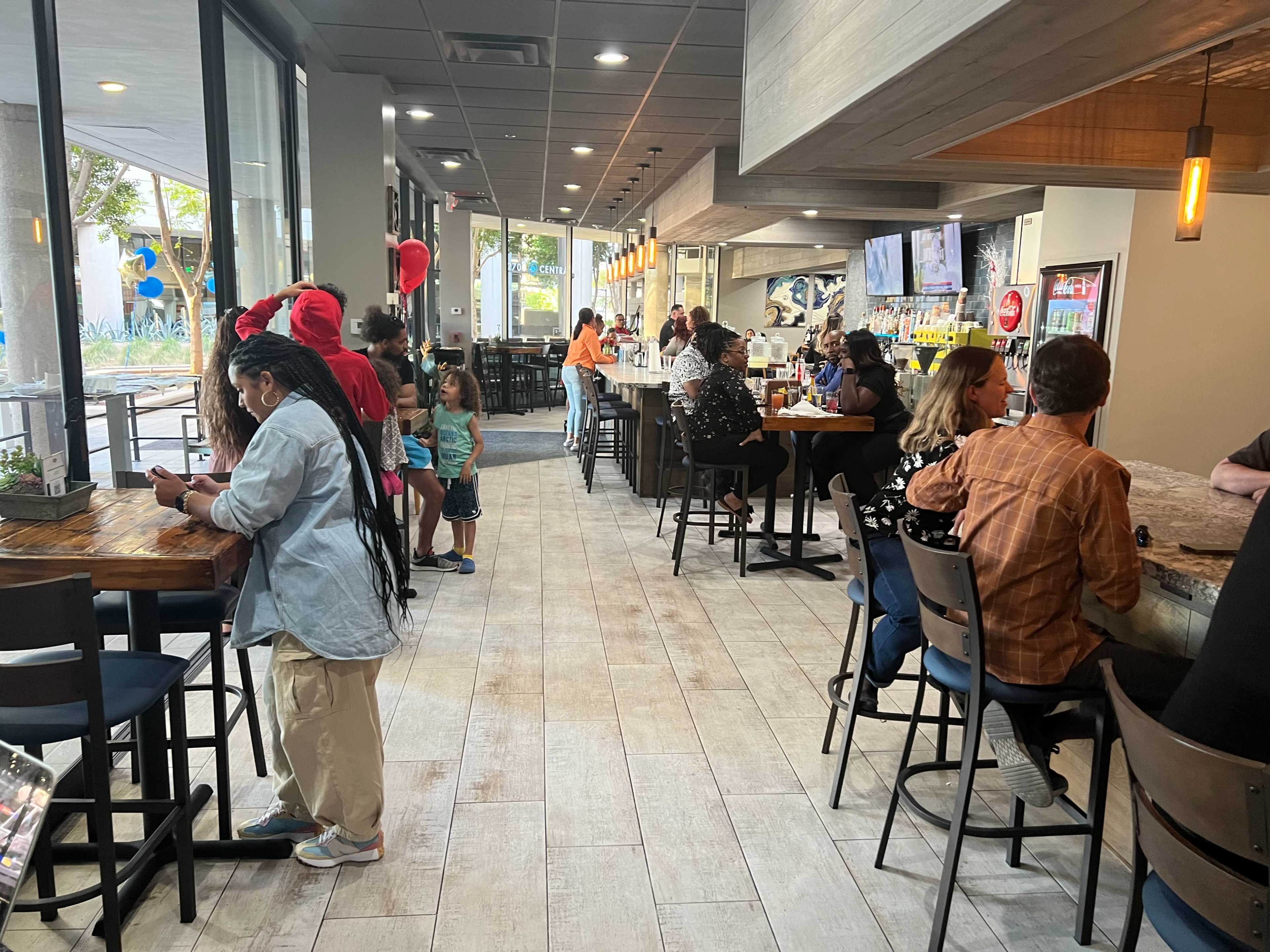 A spacious restaurant interior with patrons seated at tables and a bar, while people mingle in various areas.