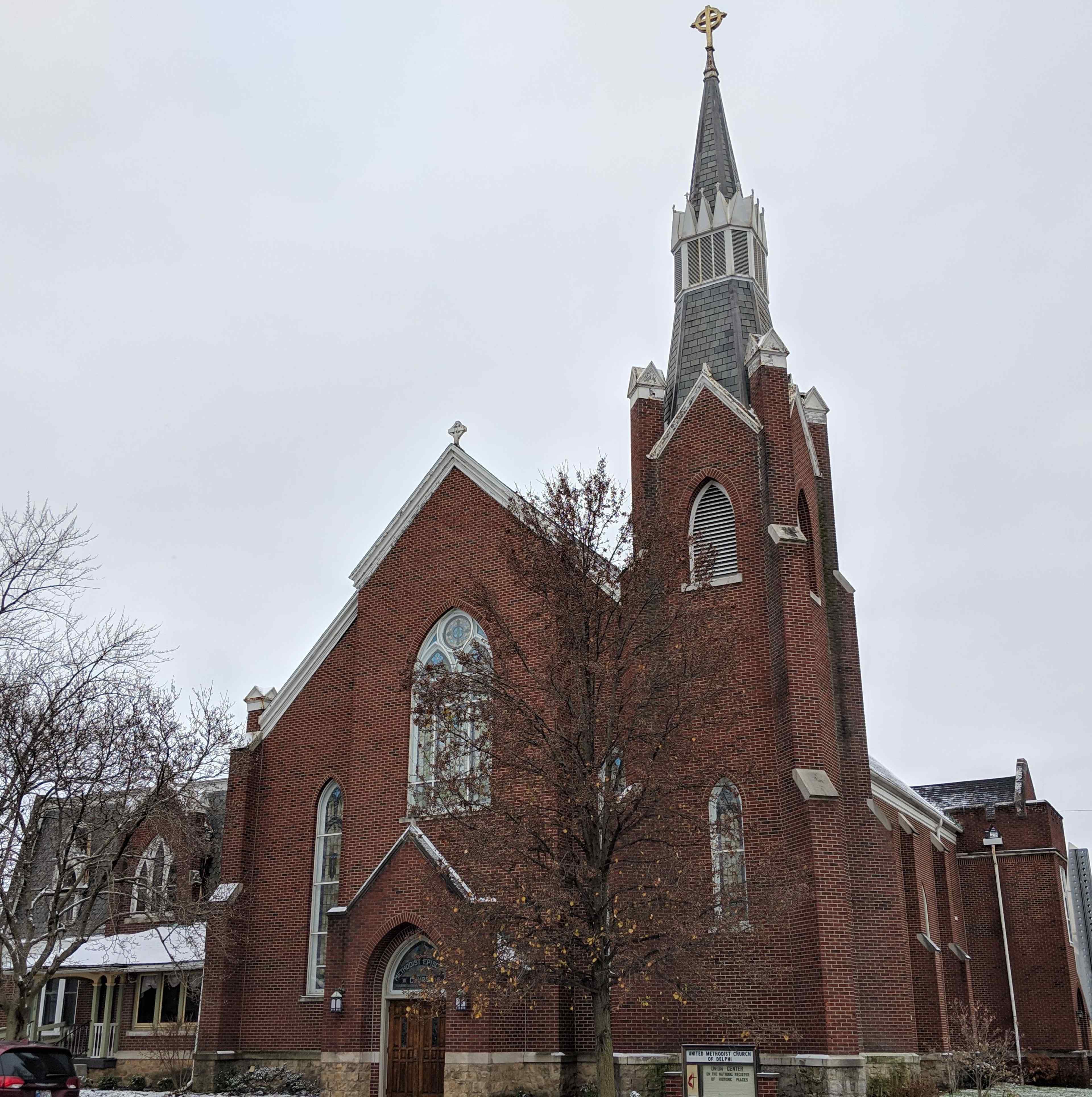 A brick church with a tall steeple and a large arched window stands beside a bare tree in a cloudy sky.