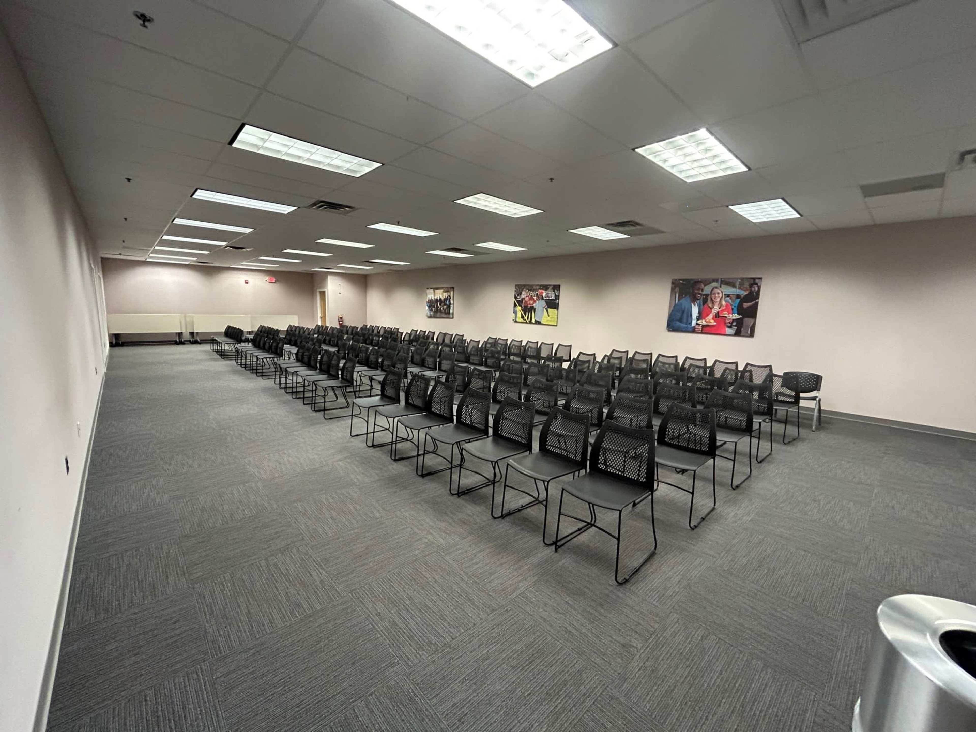 The image shows a large, empty meeting room with rows of black chairs arranged facing a blank wall, illuminated by overhead lights.