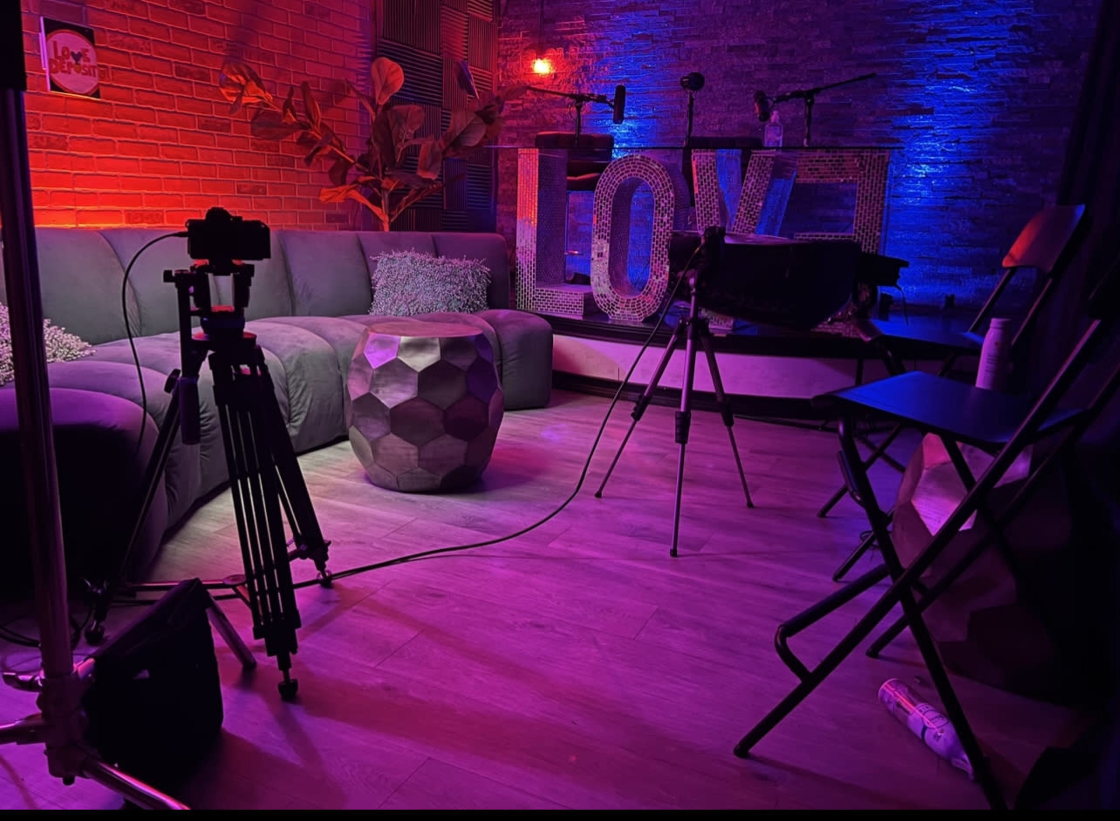 The image shows a studio setup featuring a gray sofa, a geometric table, and colorful lighting around a large "LOVE" sign.