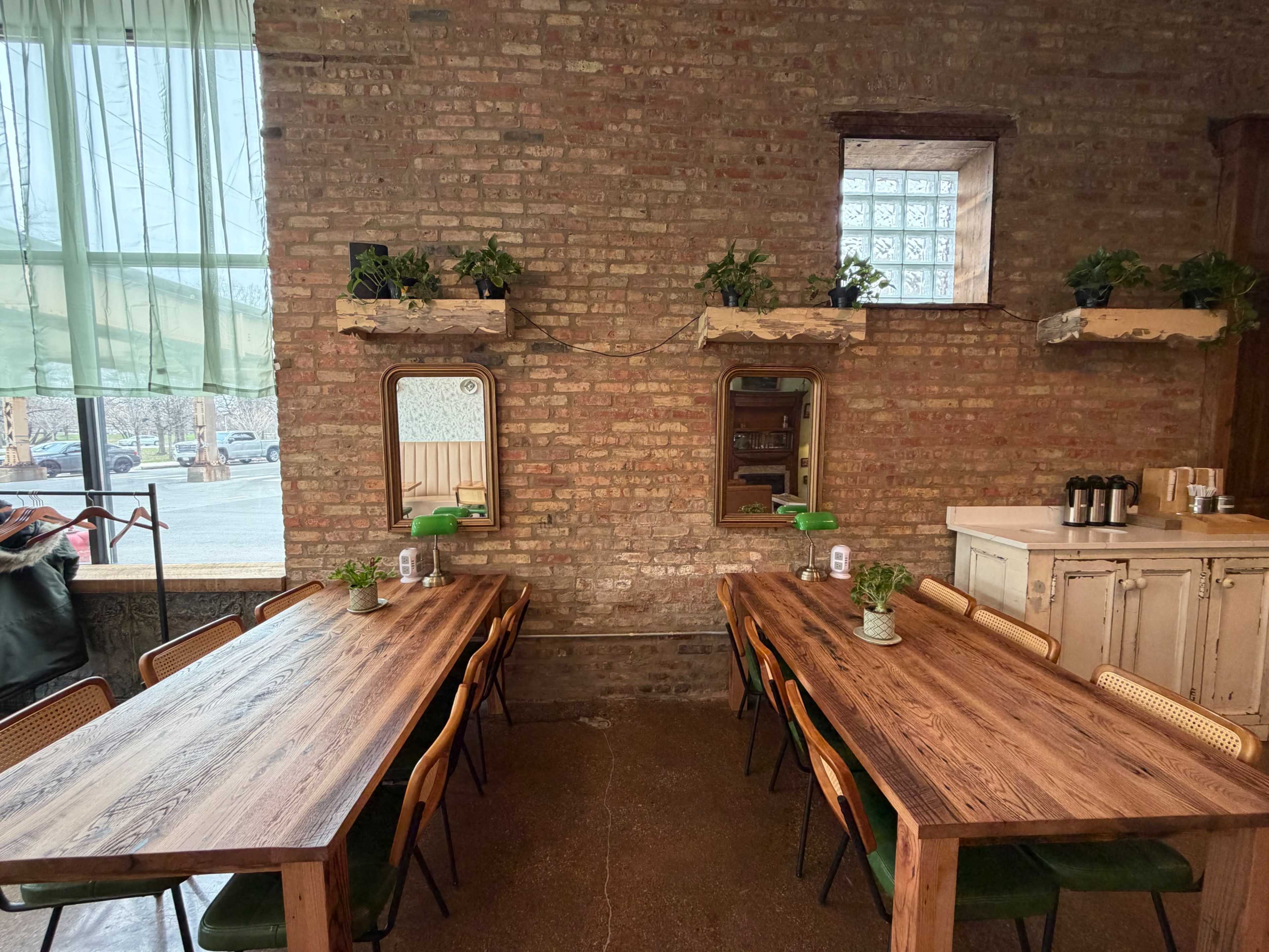 The interior of a rustic eatery features two long wooden tables, potted plants on shelves, and brick walls with large windows allowing natural light to enter.