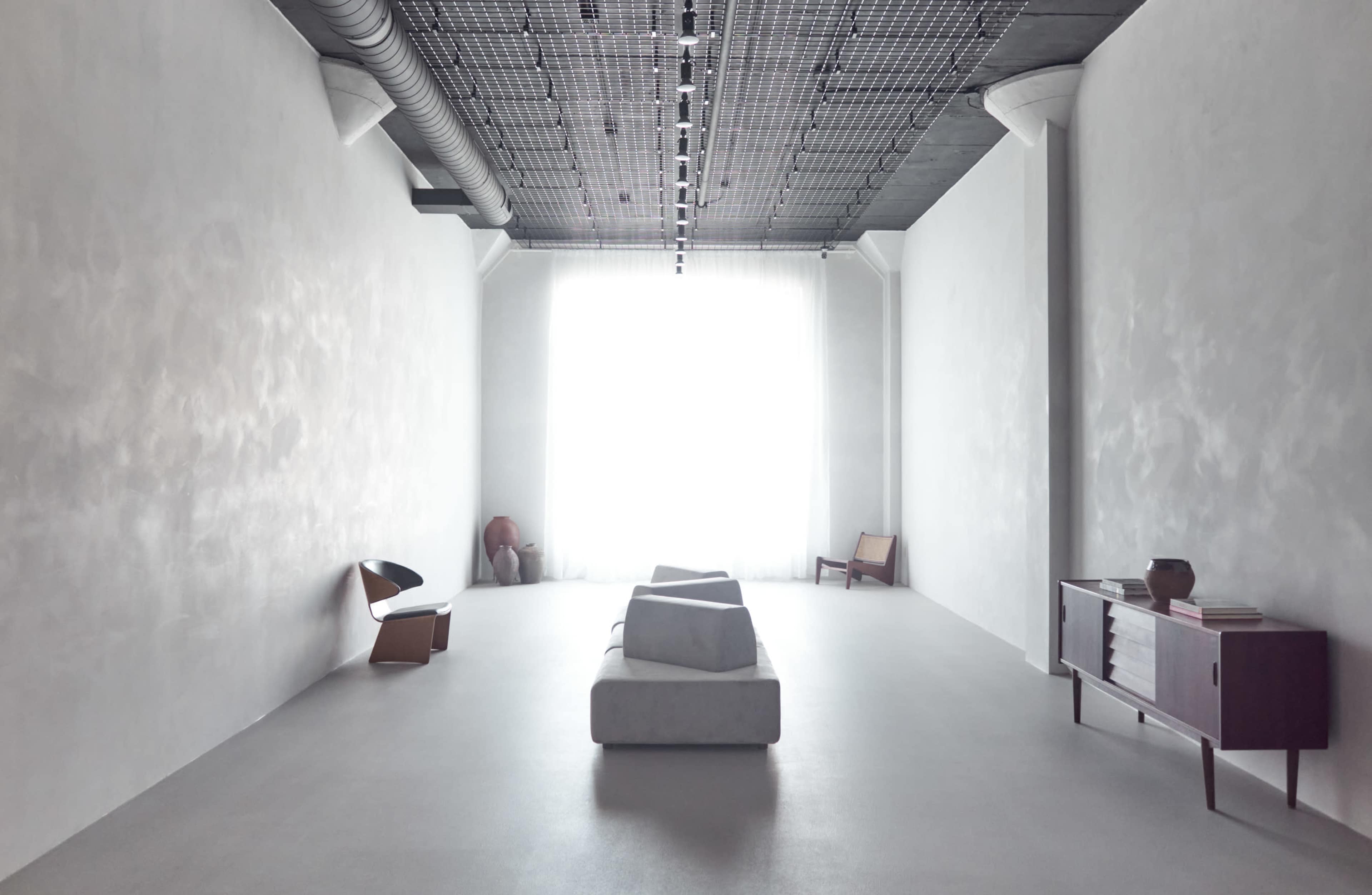 The image features a minimalist interior space with a large window, a white sofa, wooden furniture, and smooth, gray walls.