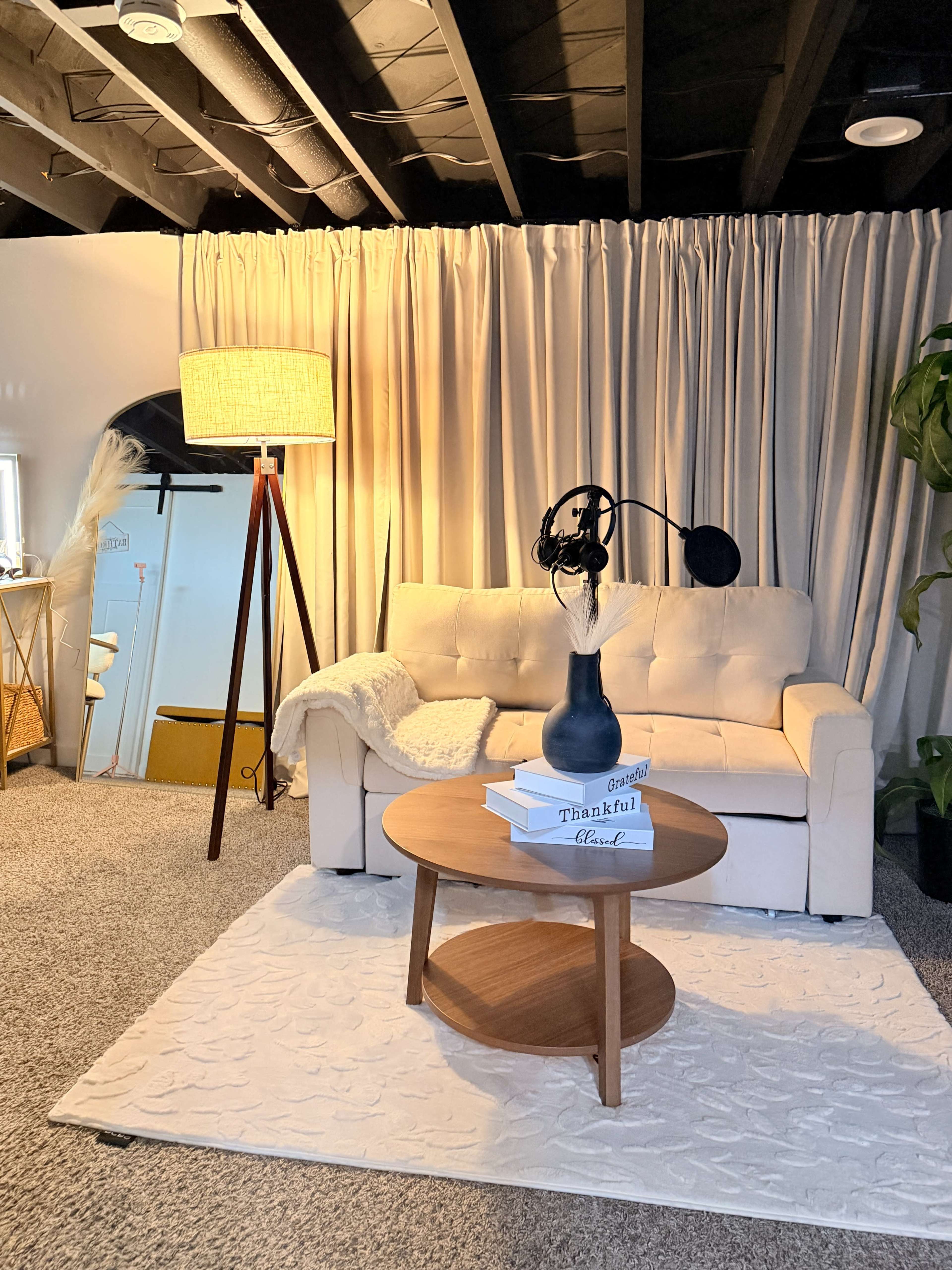 The image shows a cozy living area with a white couch, a round wooden coffee table, and a floor lamp beside a curtain backdrop.