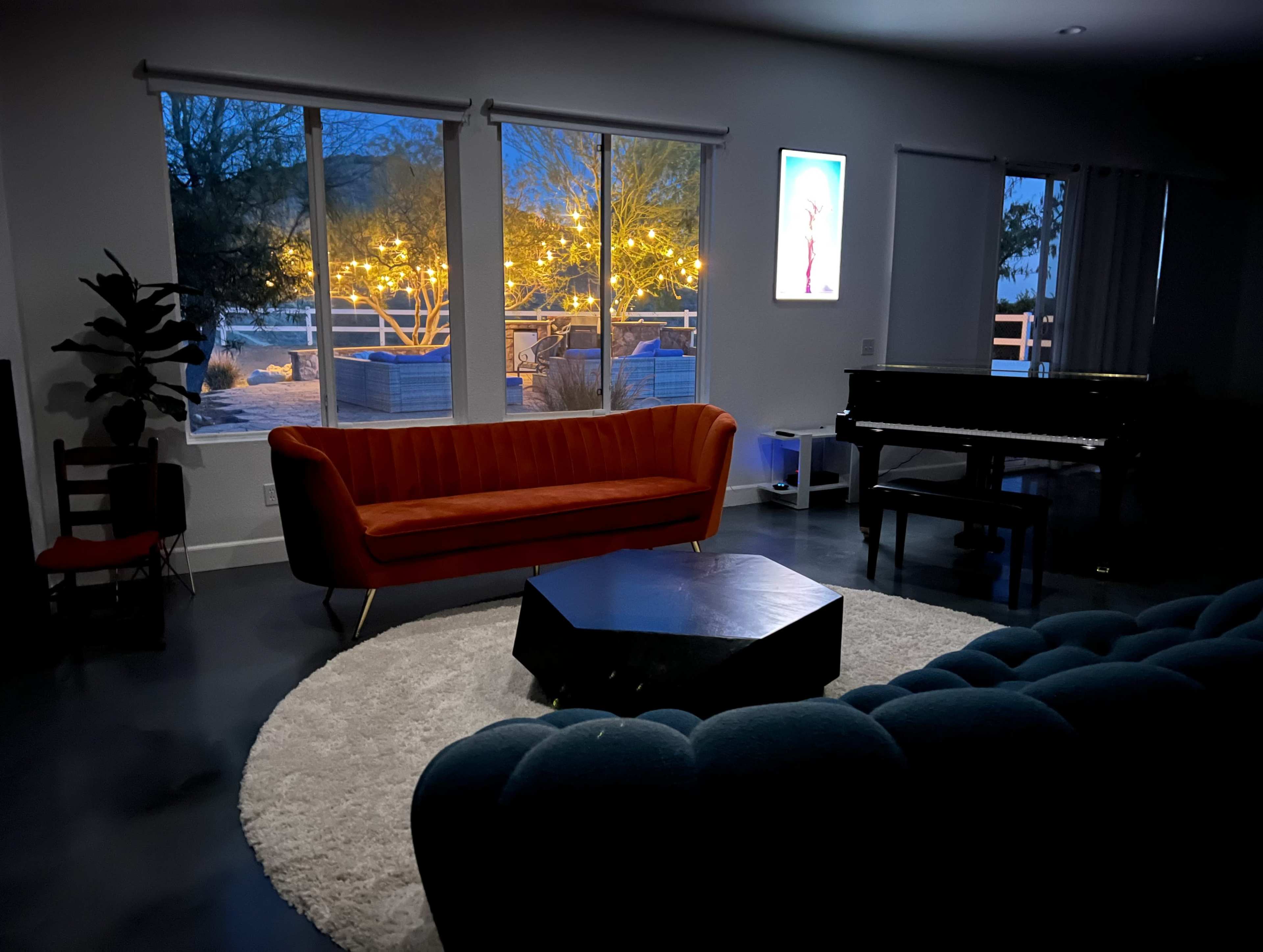 A modern living room features a bright orange sofa, a dark octagonal coffee table, a grand piano, and large windows overlooking a well-lit outdoor area.