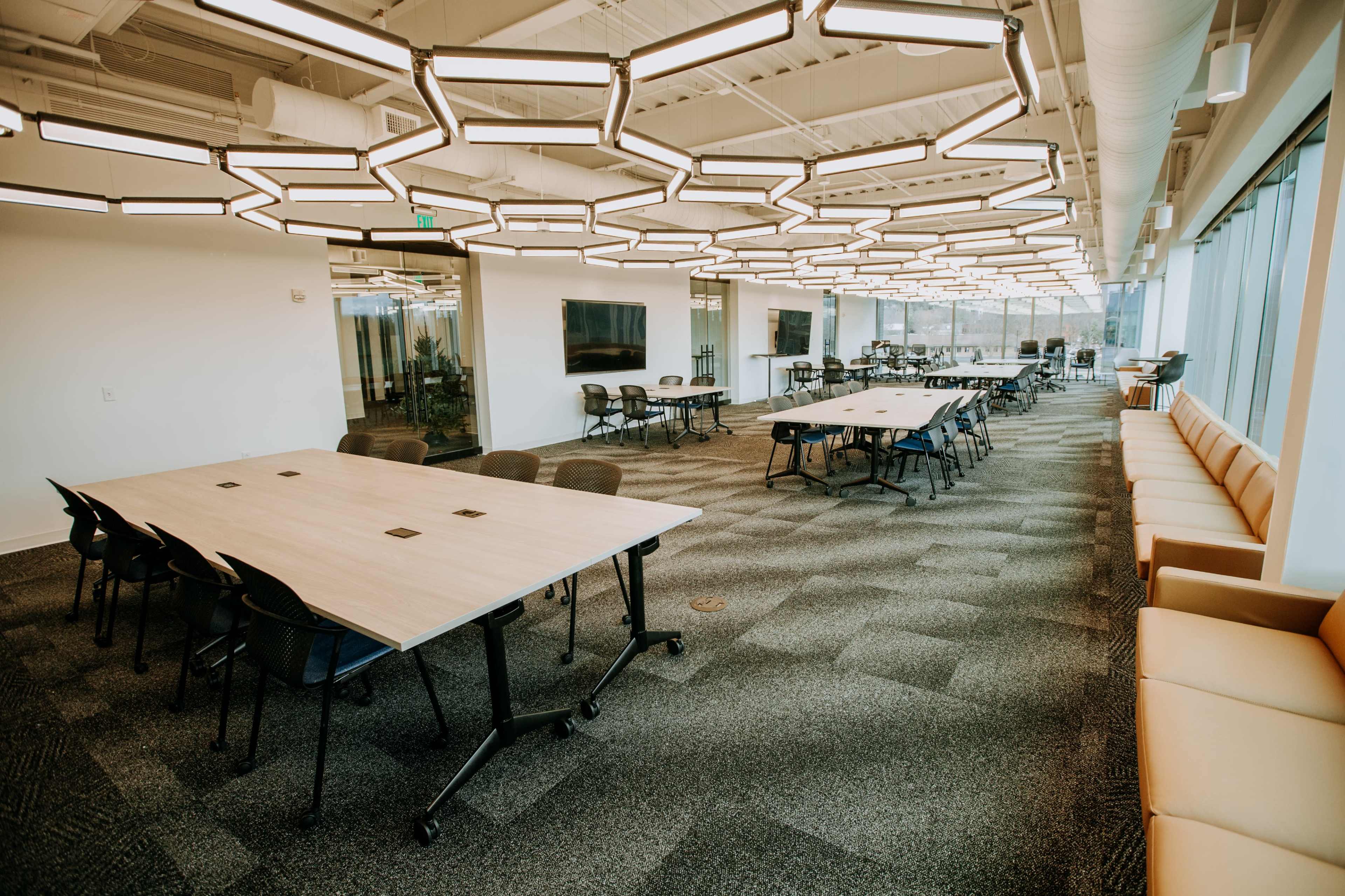 The image shows a spacious, modern office interior with multiple large tables, chairs, and an area featuring a beige couch.