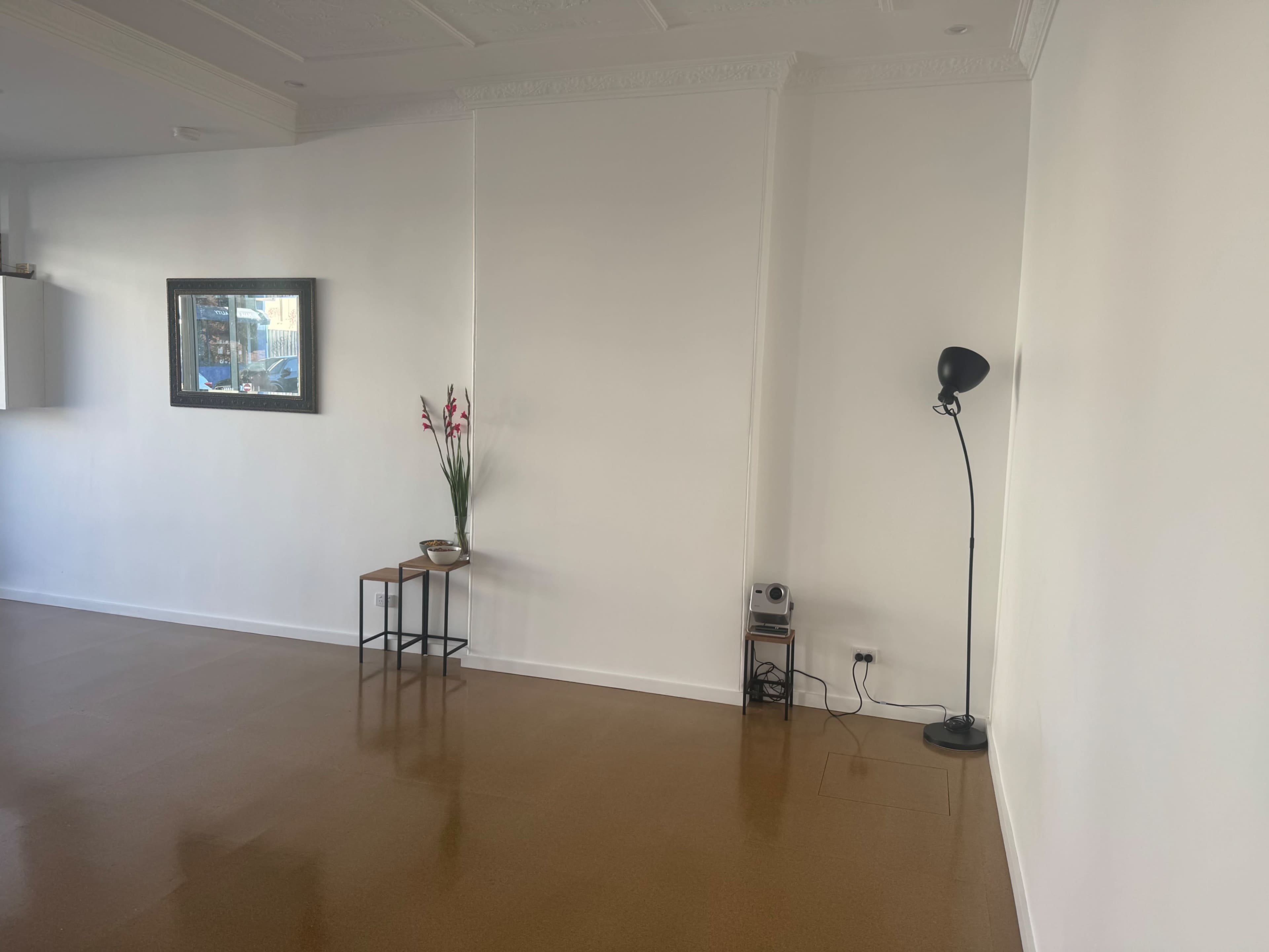 The image shows a minimalist room featuring a small table with a vase of flowers, a framed mirror on the wall, and a floor lamp in the corner.