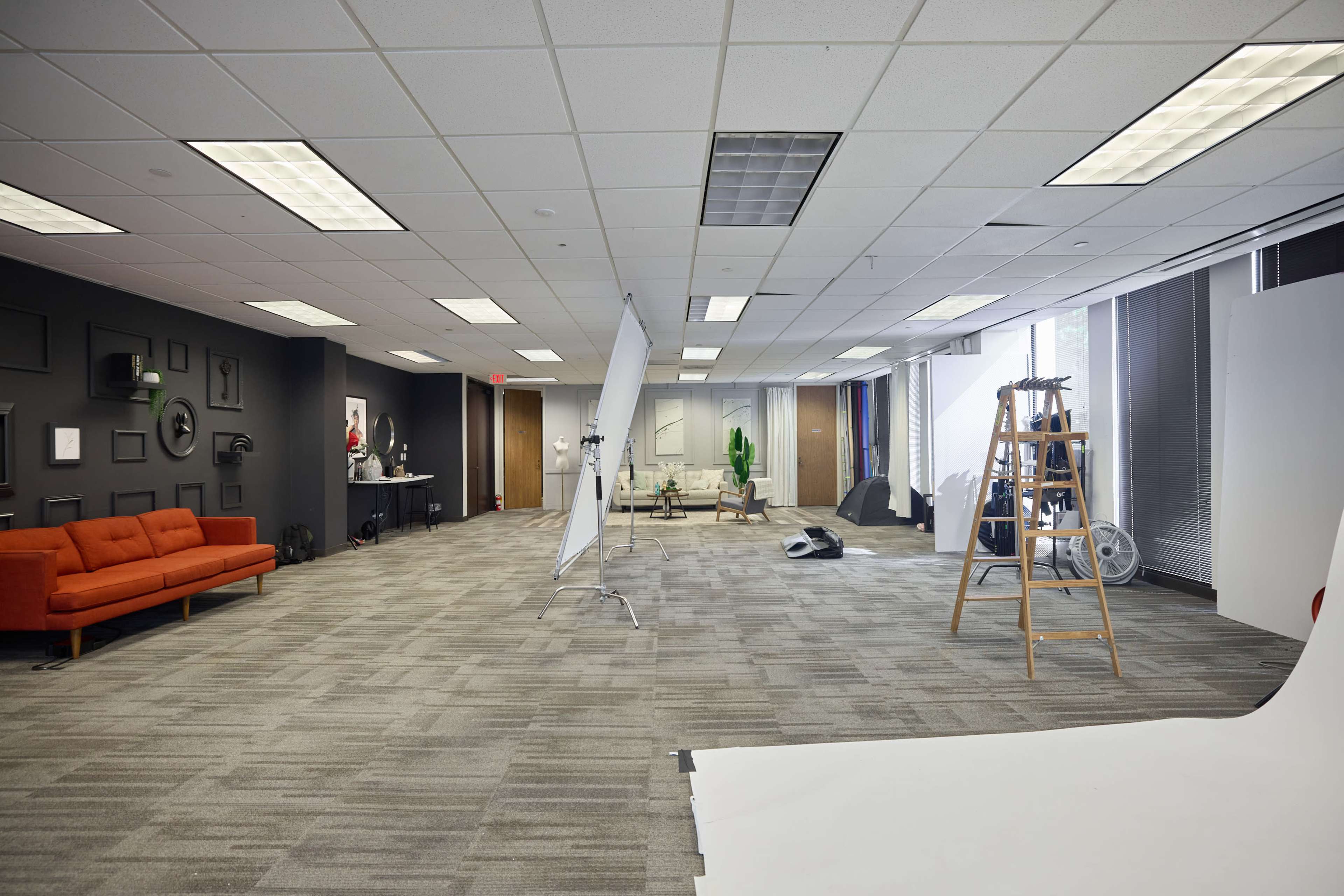 An empty studio space features a red sofa, a light stand, a ladder, and a white backdrop set up near large windows.