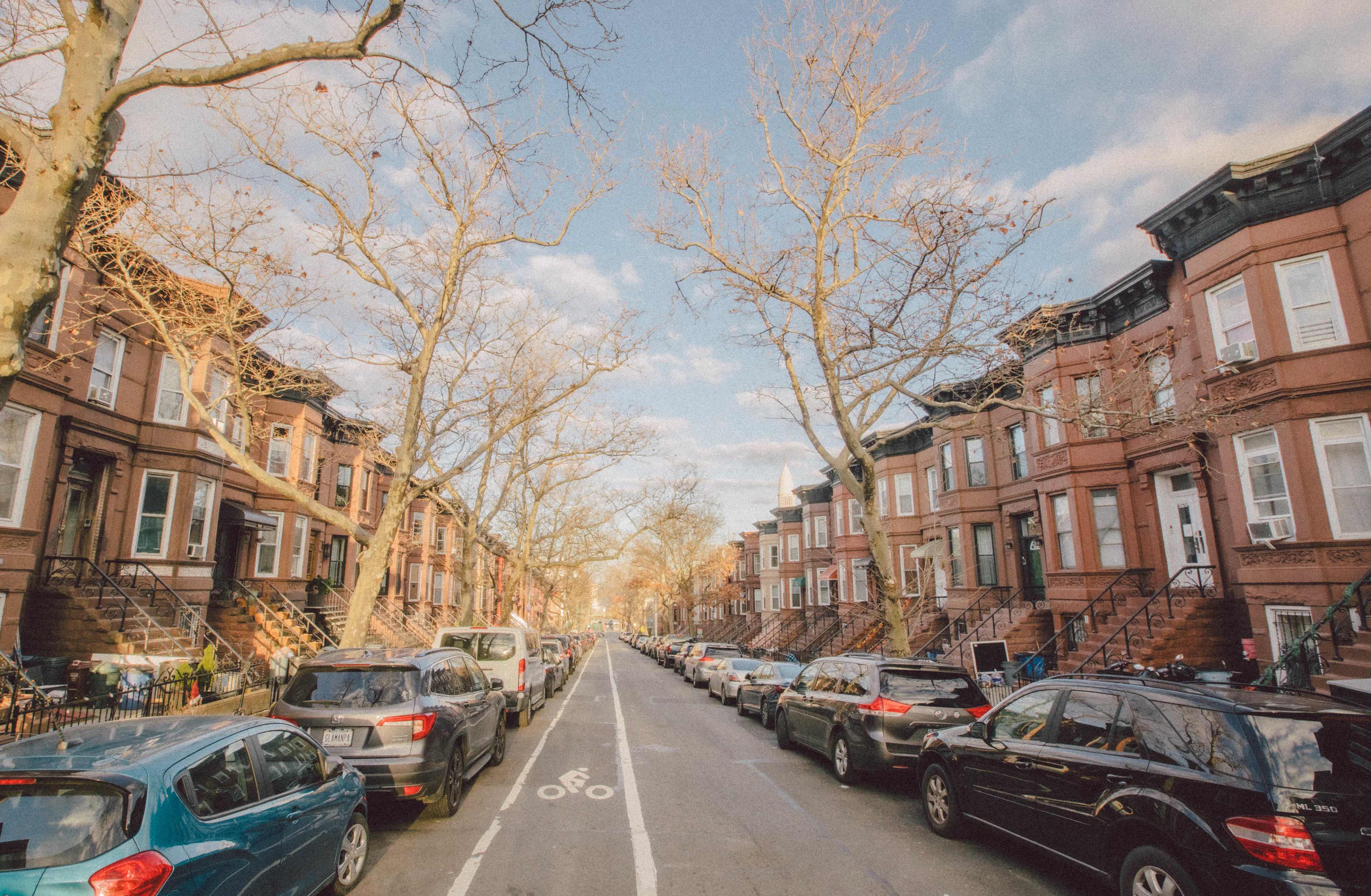 A tree-lined street features brownstone buildings on either side, with parked cars lining the road.