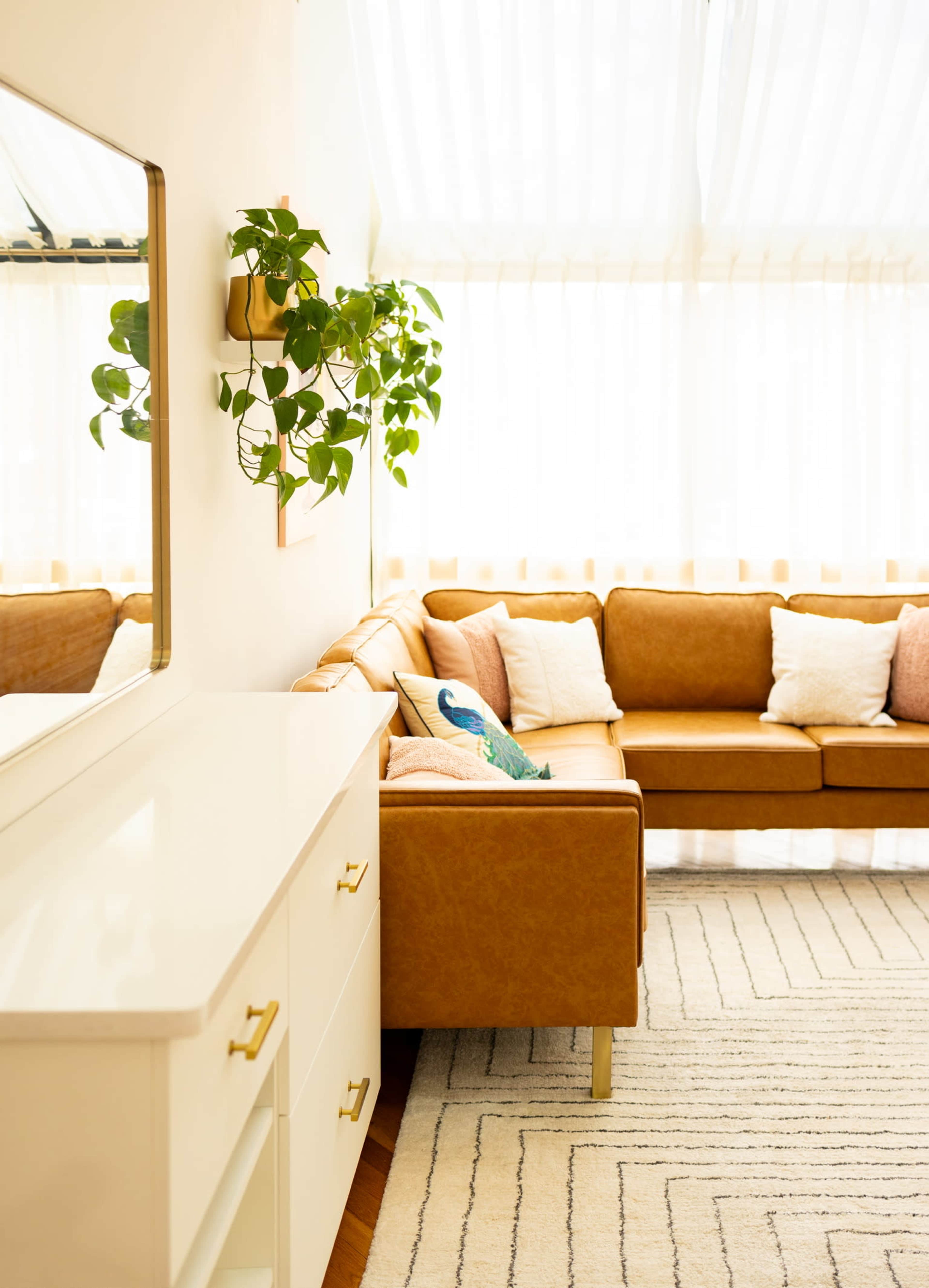 A well-lit living room features a brown leather sofa, a white dresser, and a plant hanging on the wall.
