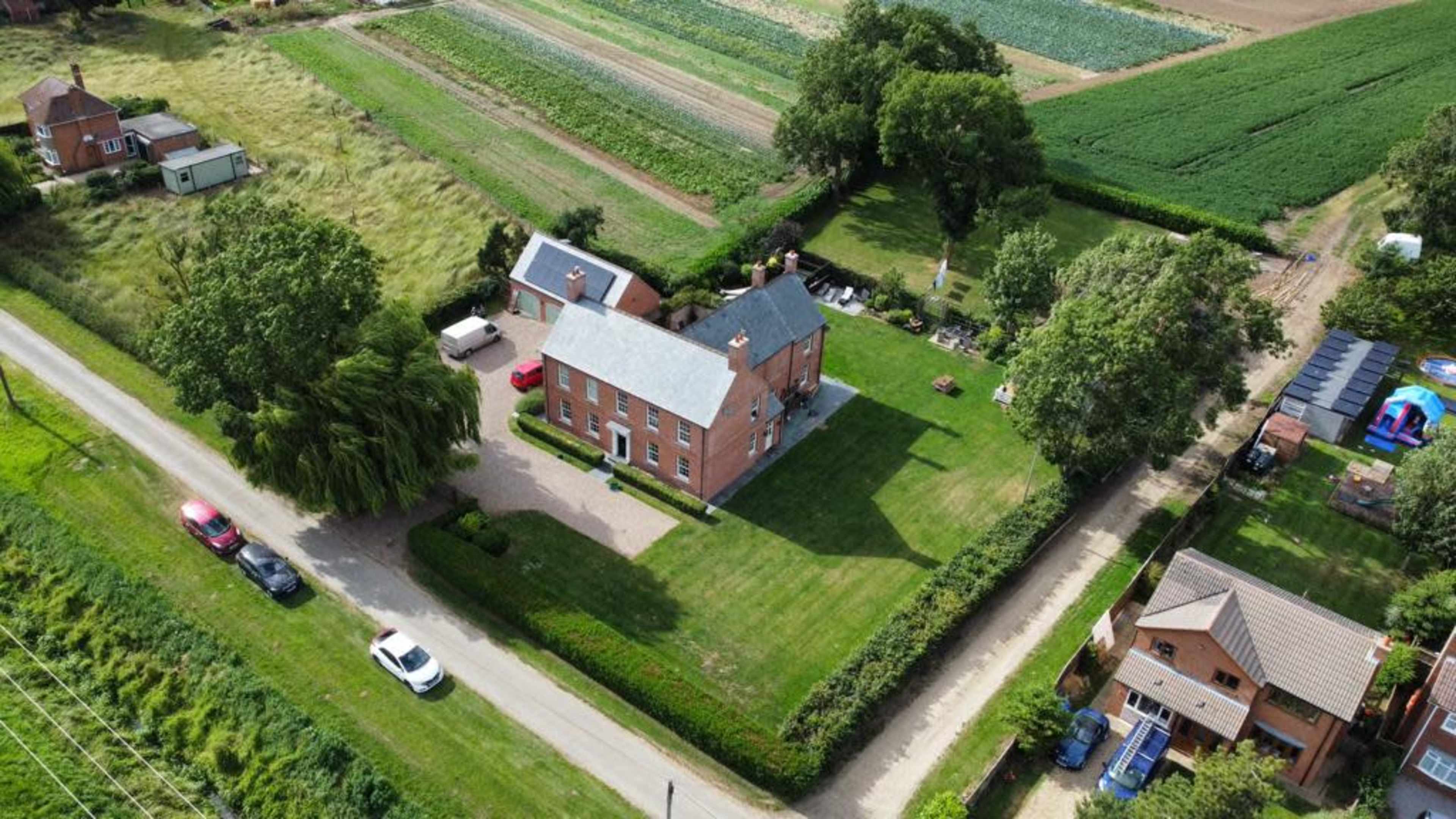 A large brick house with a well-maintained lawn, surrounded by greenery and agricultural fields.