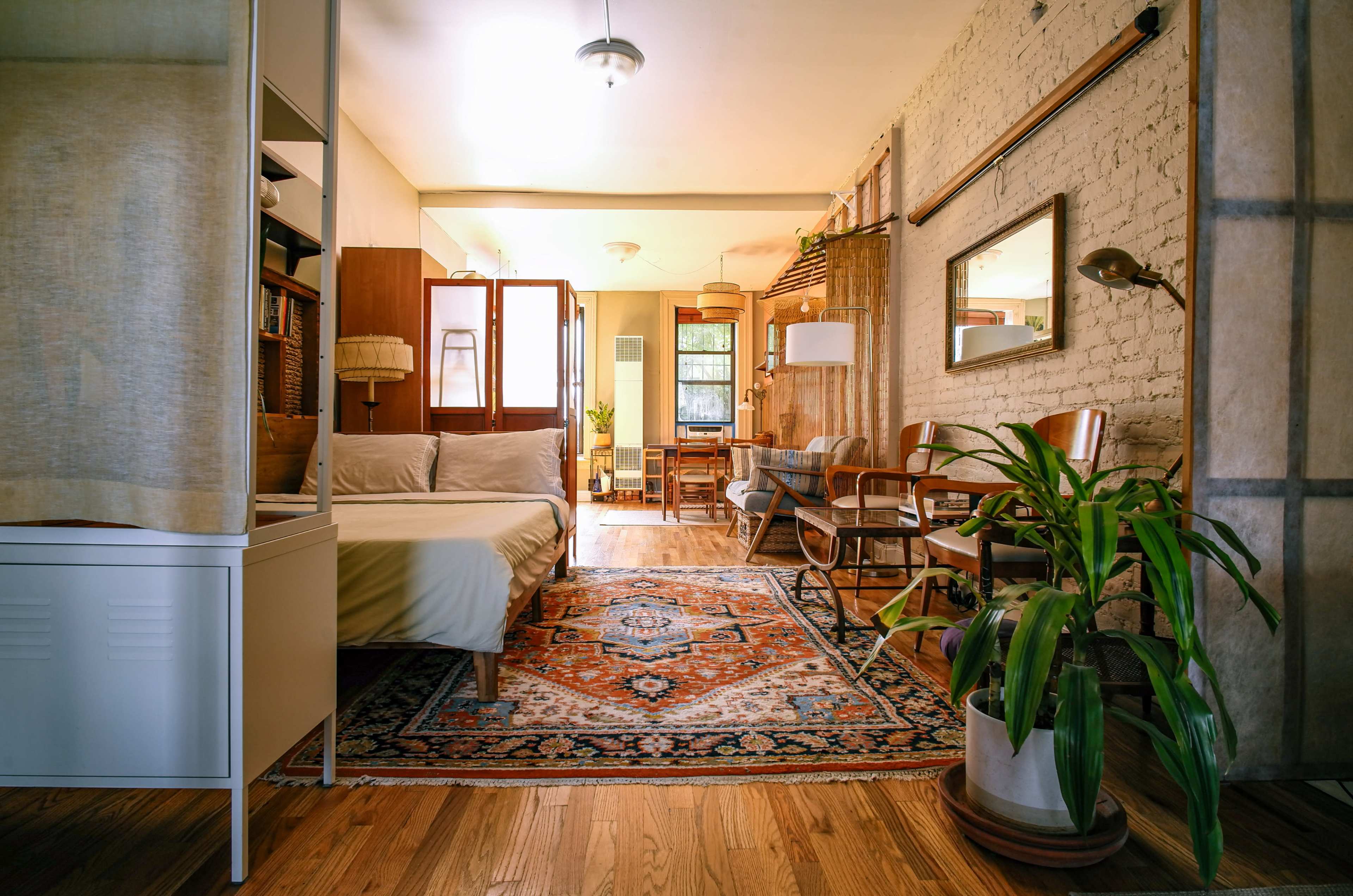 The image shows a cozy, well-furnished living space featuring a bed, a dining area with chairs, a decorative rug, and plants against a backdrop of exposed brick walls.