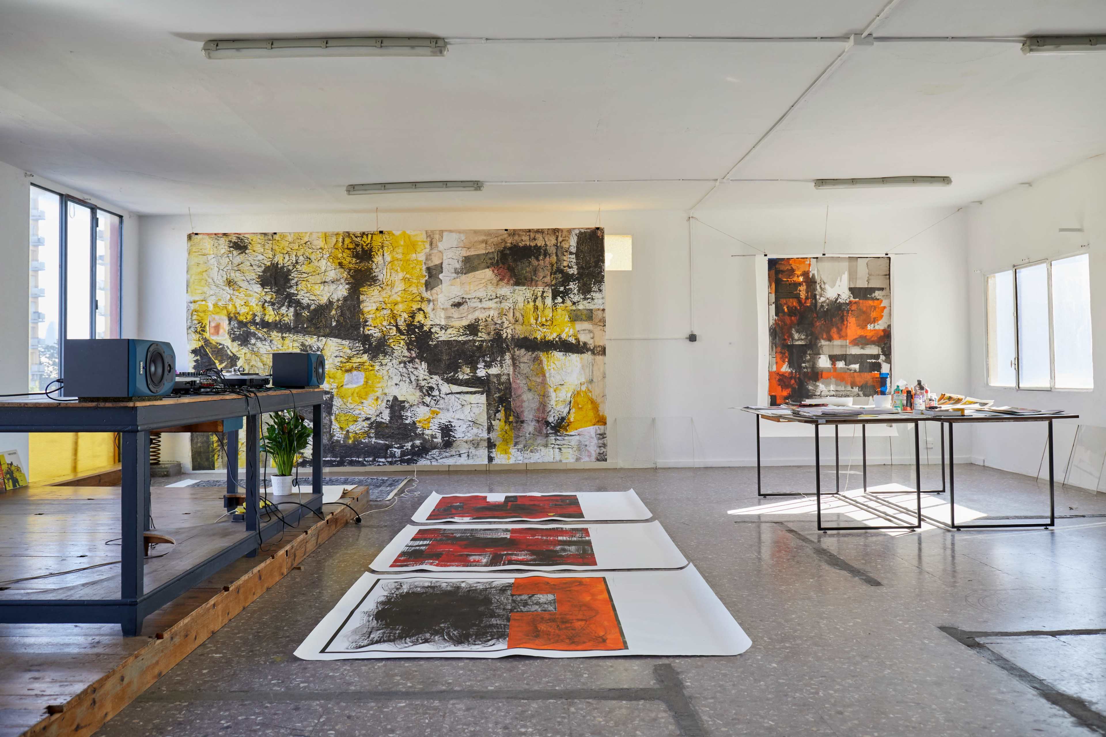 An art studio with large abstract paintings on the walls, a table with art supplies, and several canvases laid out on the floor.