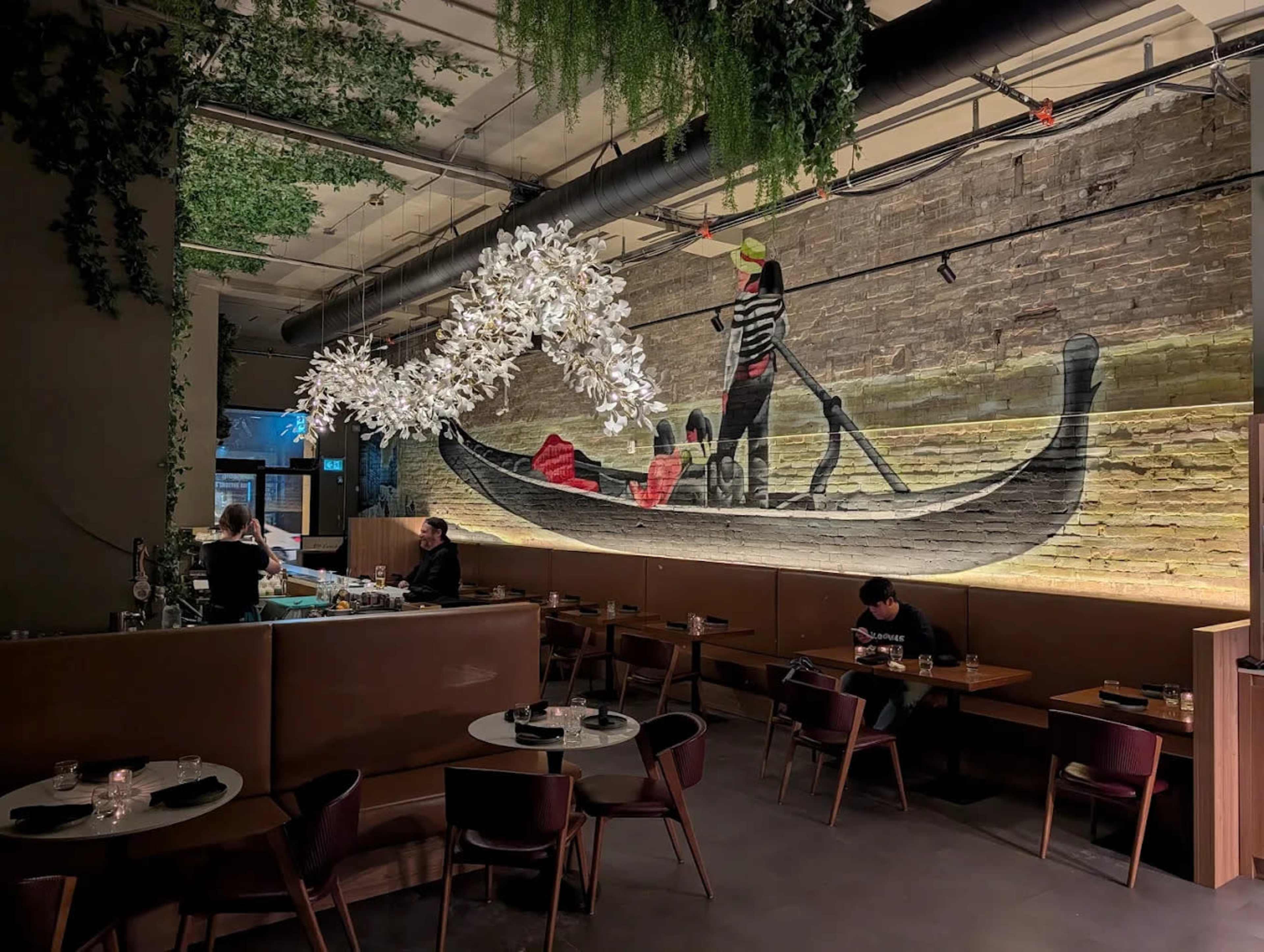 The interior of a restaurant features a large mural of a gondola scene, with diners seated at tables below.