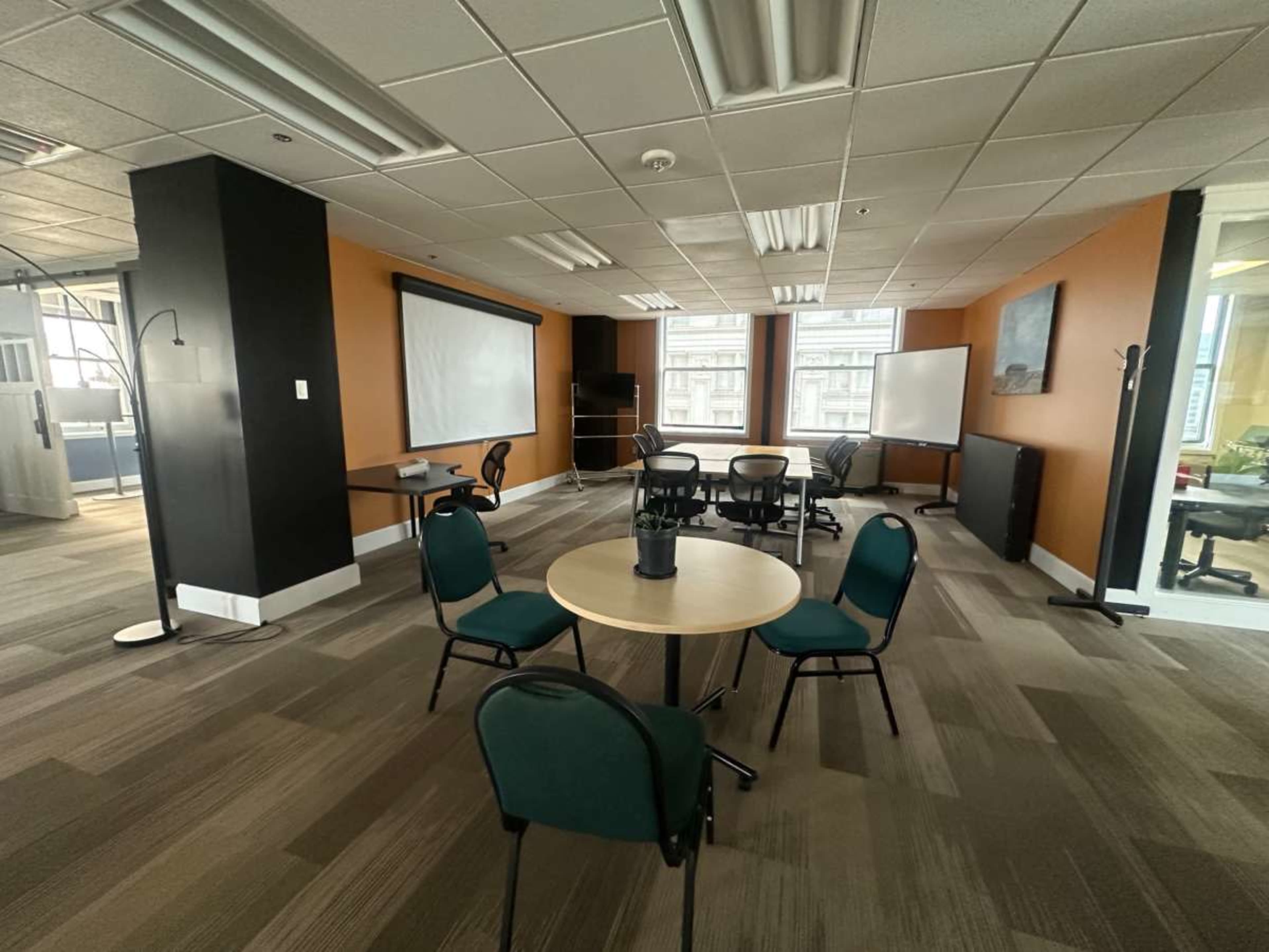 The image shows an office space with a circular table surrounded by chairs, a blank projector screen, and several workstations against orange and black walls.