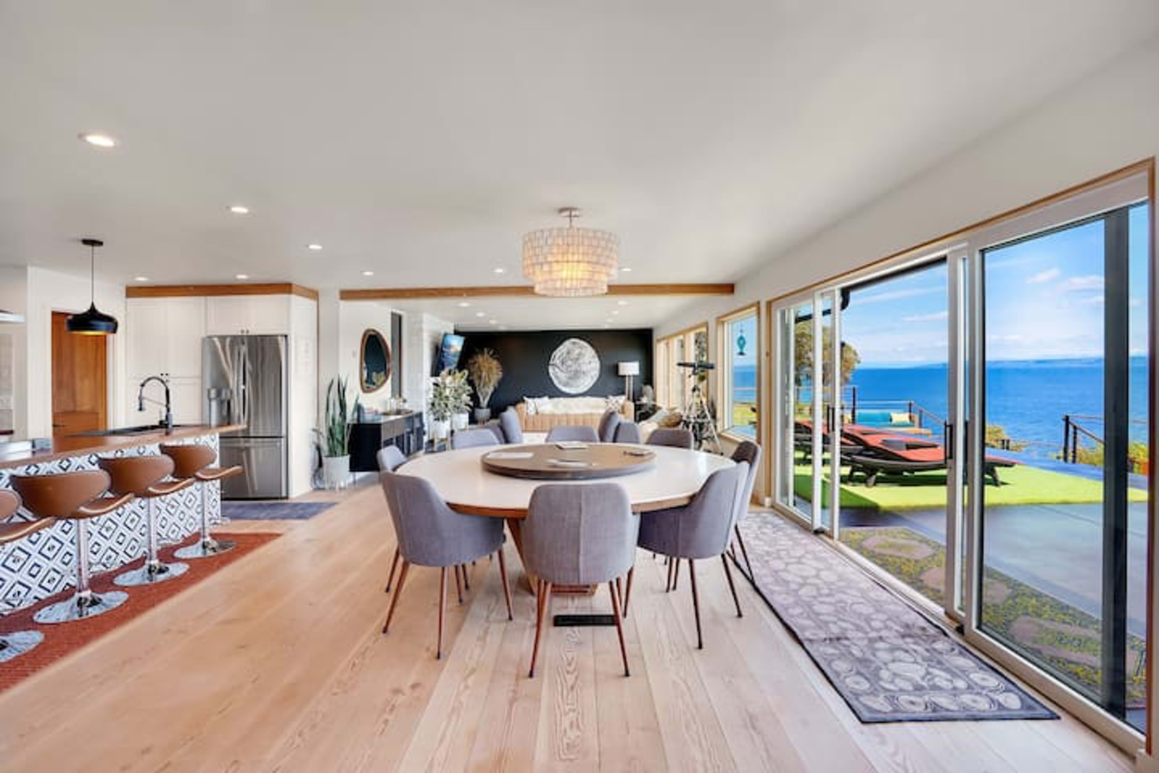A bright, open-concept dining and living area with large windows overlooking a water view, featuring a round dining table surrounded by gray chairs and a modern kitchen.