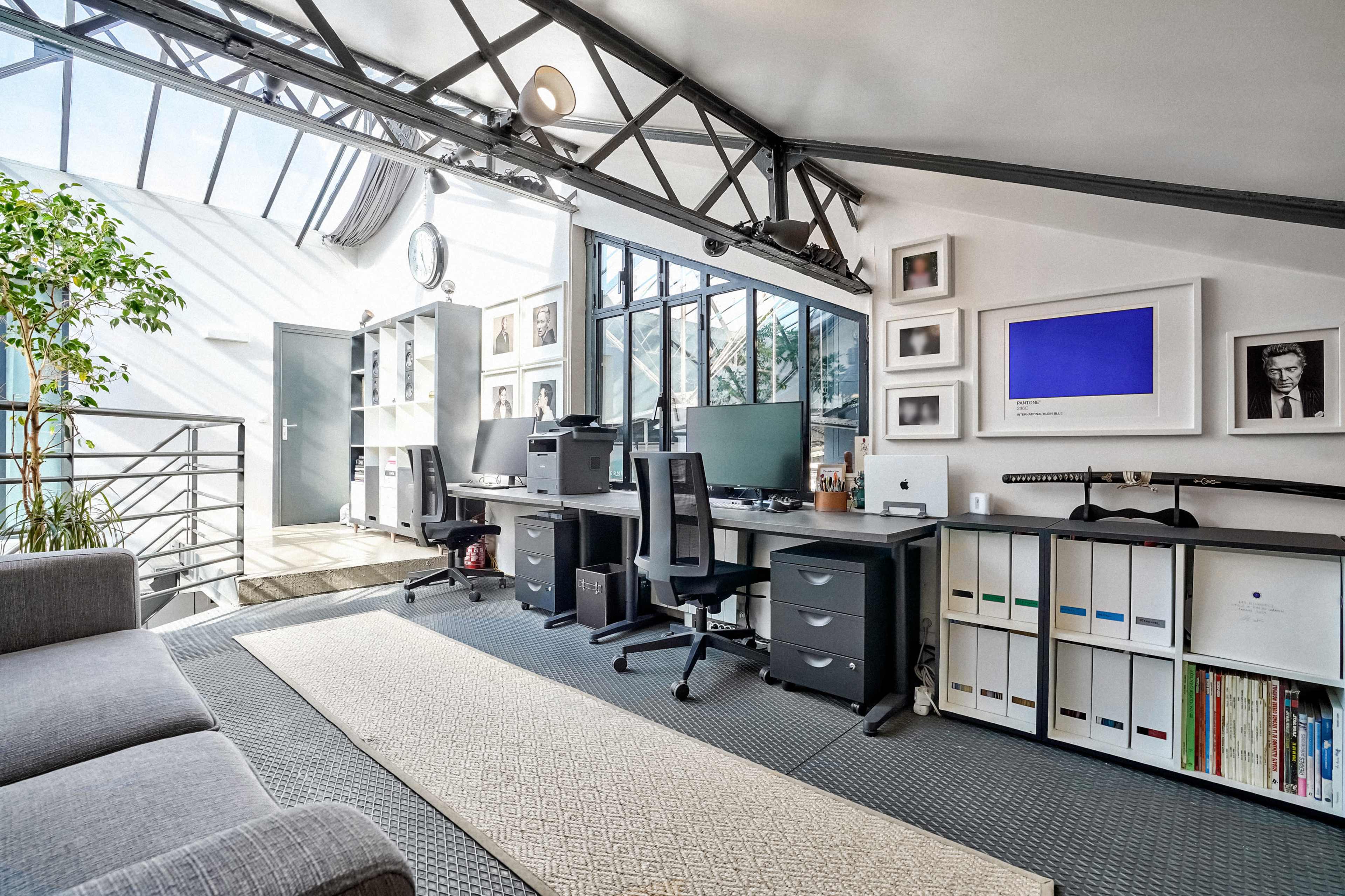 The image shows a modern home office with large windows, two desks equipped with computers, and neatly organized shelves.