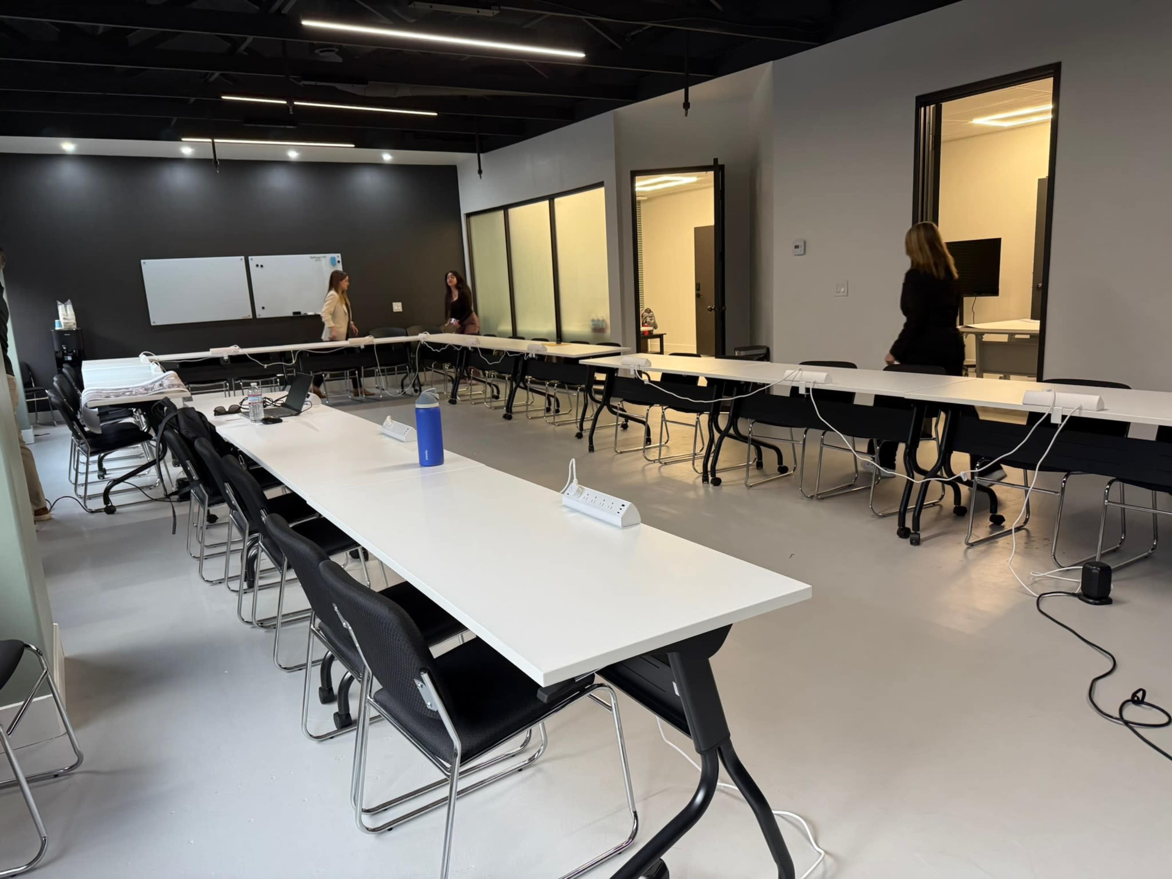 A spacious, modern meeting room features long tables arranged in a U-shape, with several people moving about in the background.