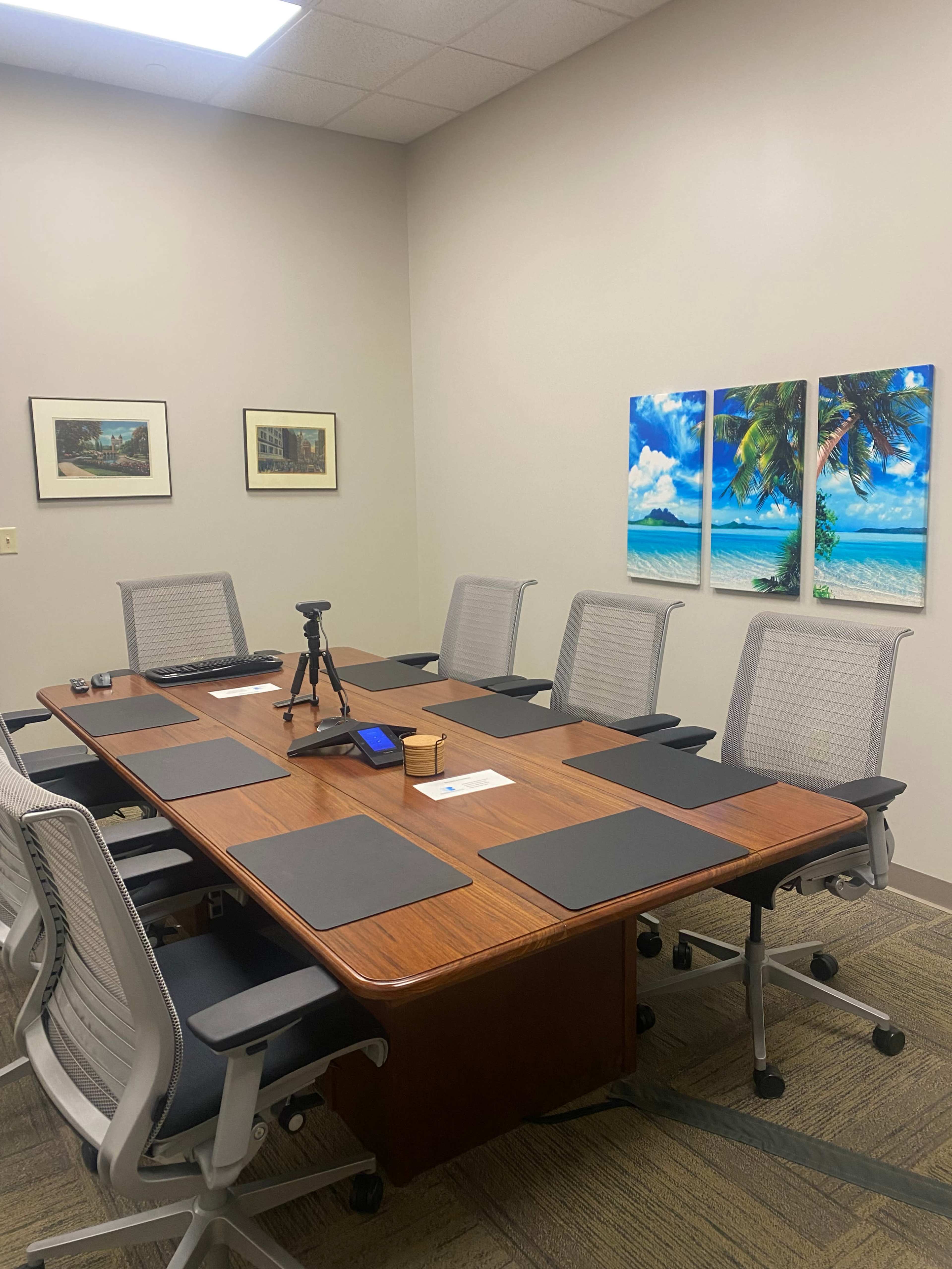 A conference room features a large wooden table surrounded by six ergonomic chairs, with a camera setup and tropical-themed artwork on the walls.