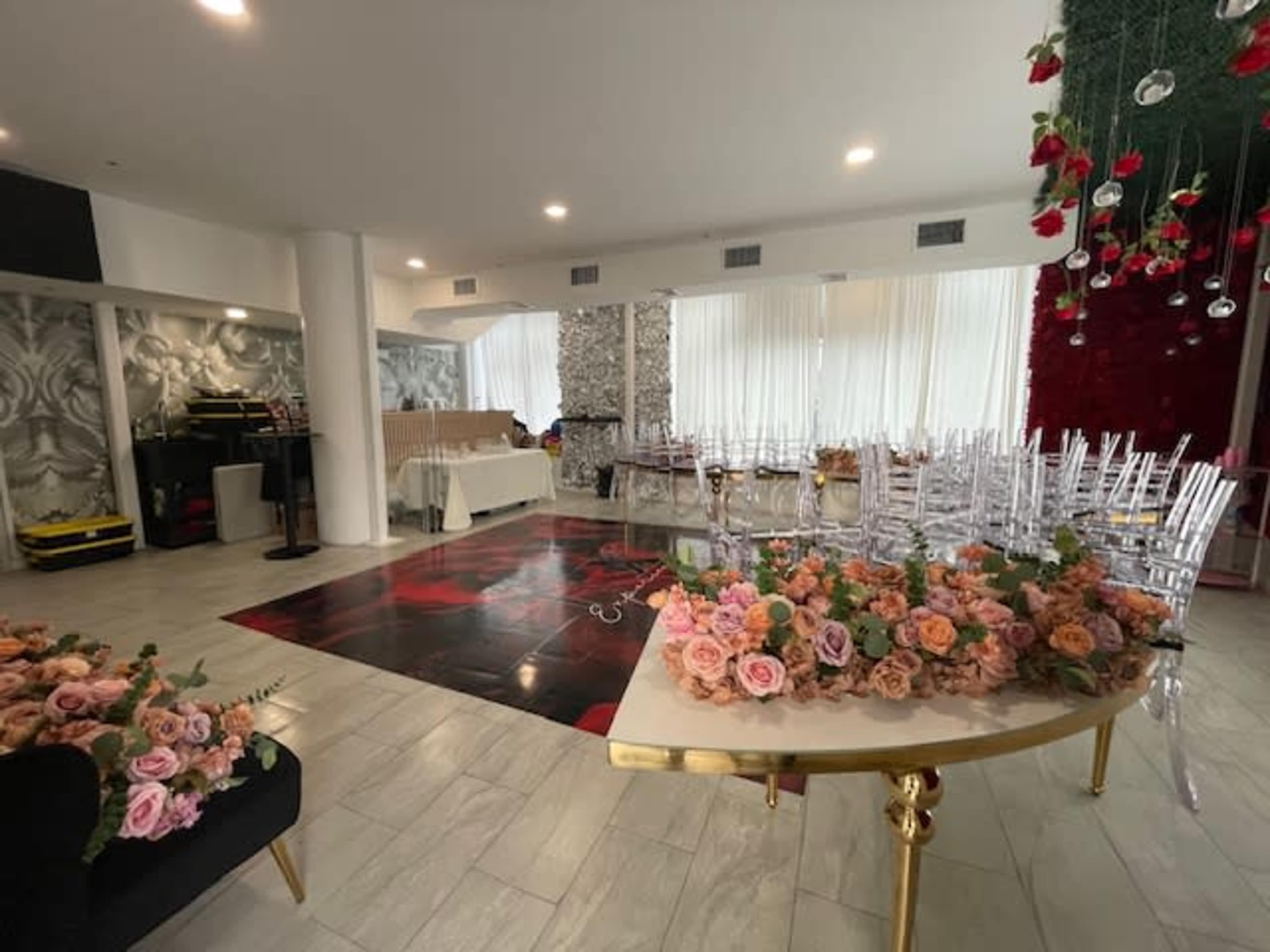 The image shows a decorated event space featuring a large table with floral arrangements, transparent chairs, and a patterned floor.