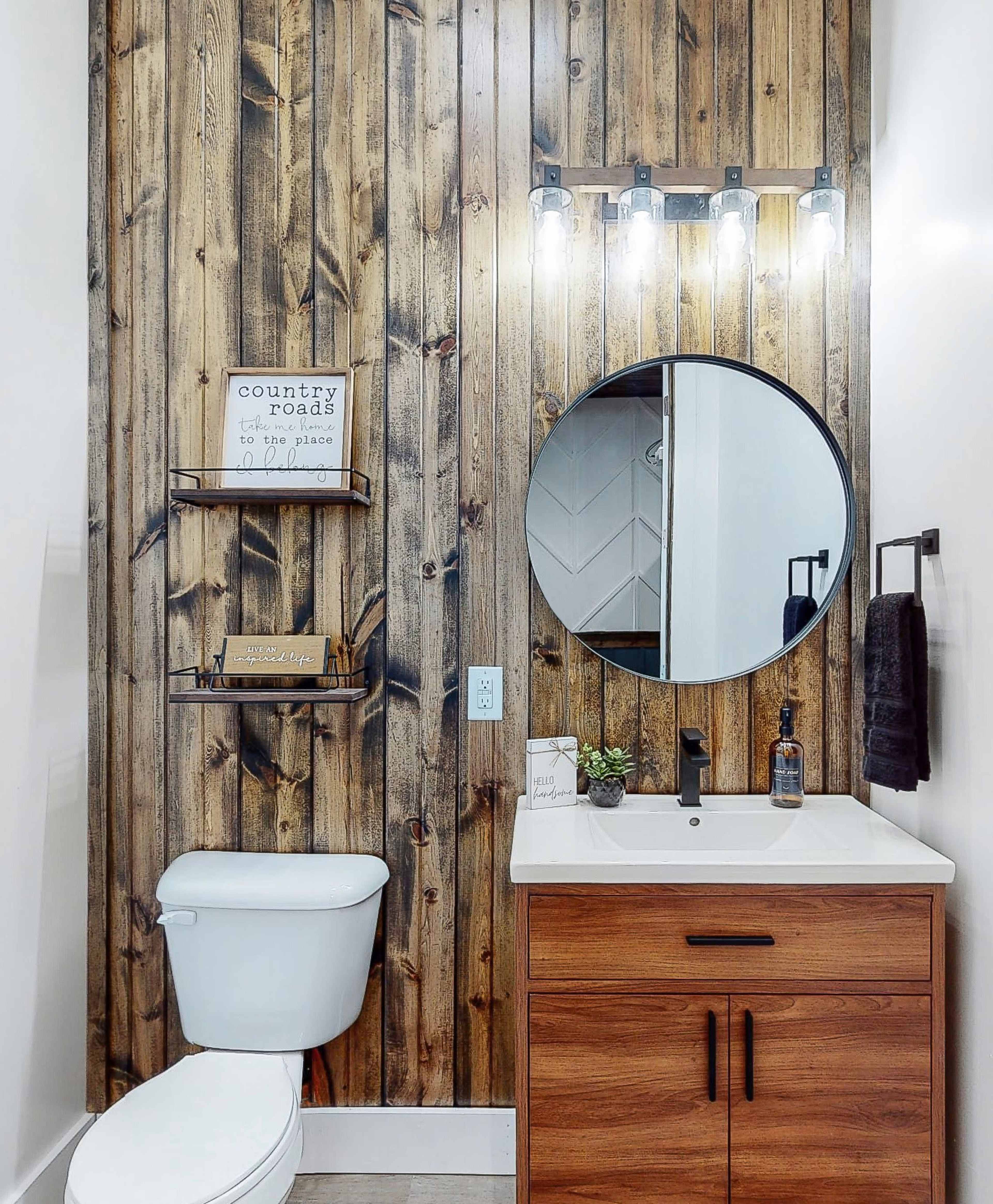 A modern bathroom featuring a wooden accent wall, a circular mirror, a white sink with a wooden vanity, and a toilet beside floating shelves.