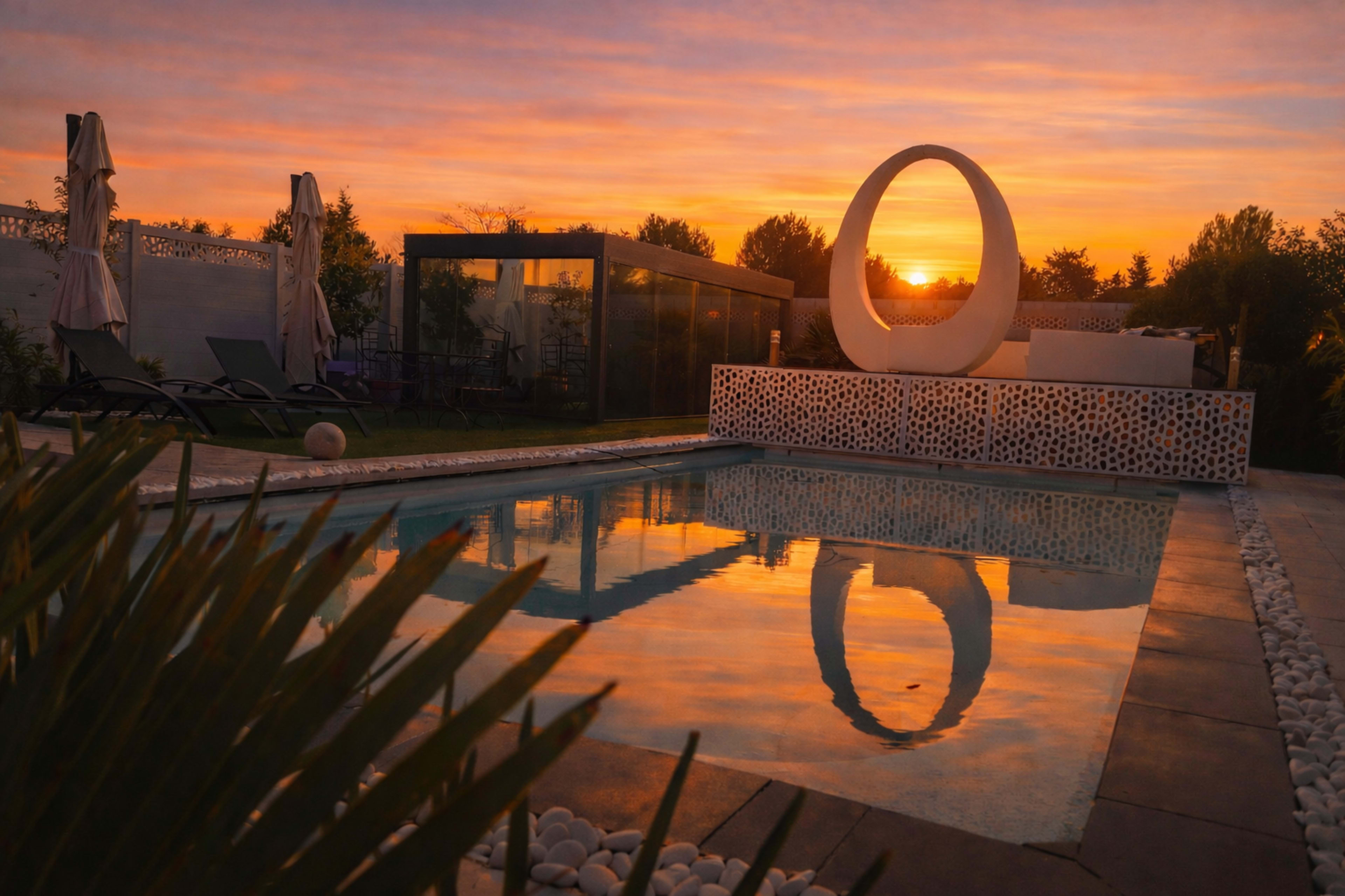The scene features a swimming pool reflecting a decorative sculpture at sunset, with lounge chairs and umbrellas in the background.