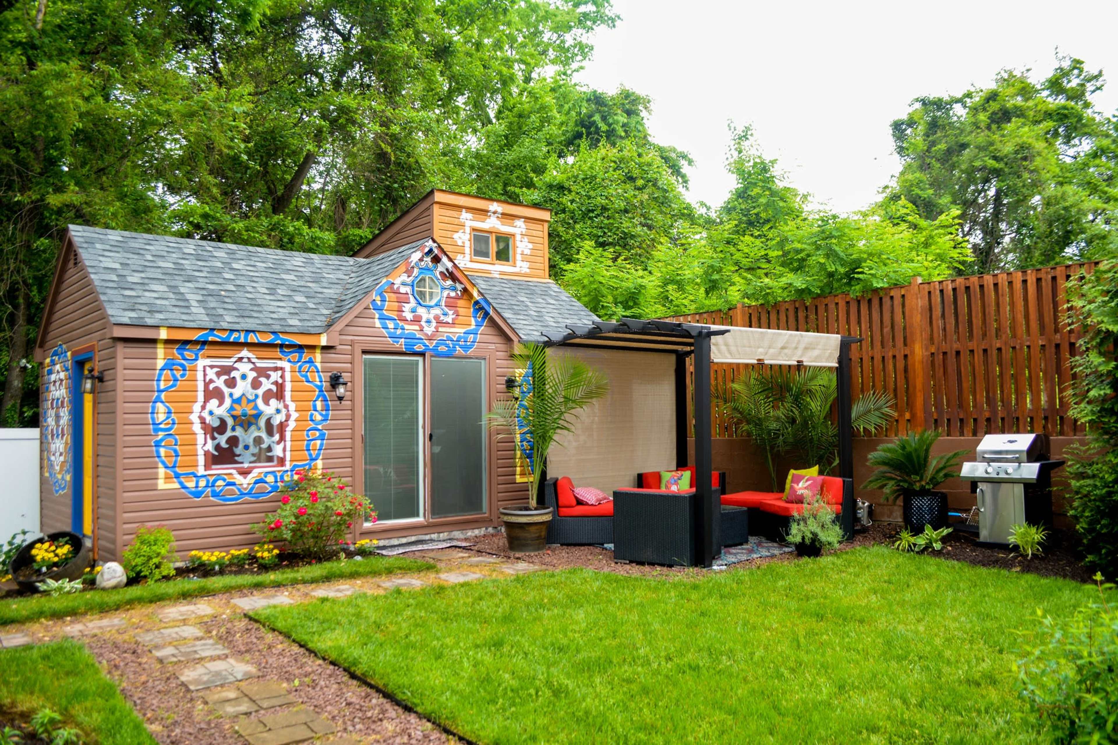 A small, colorful garden shed surrounded by greenery, with a patio area featuring red seating and a barbecue grill.