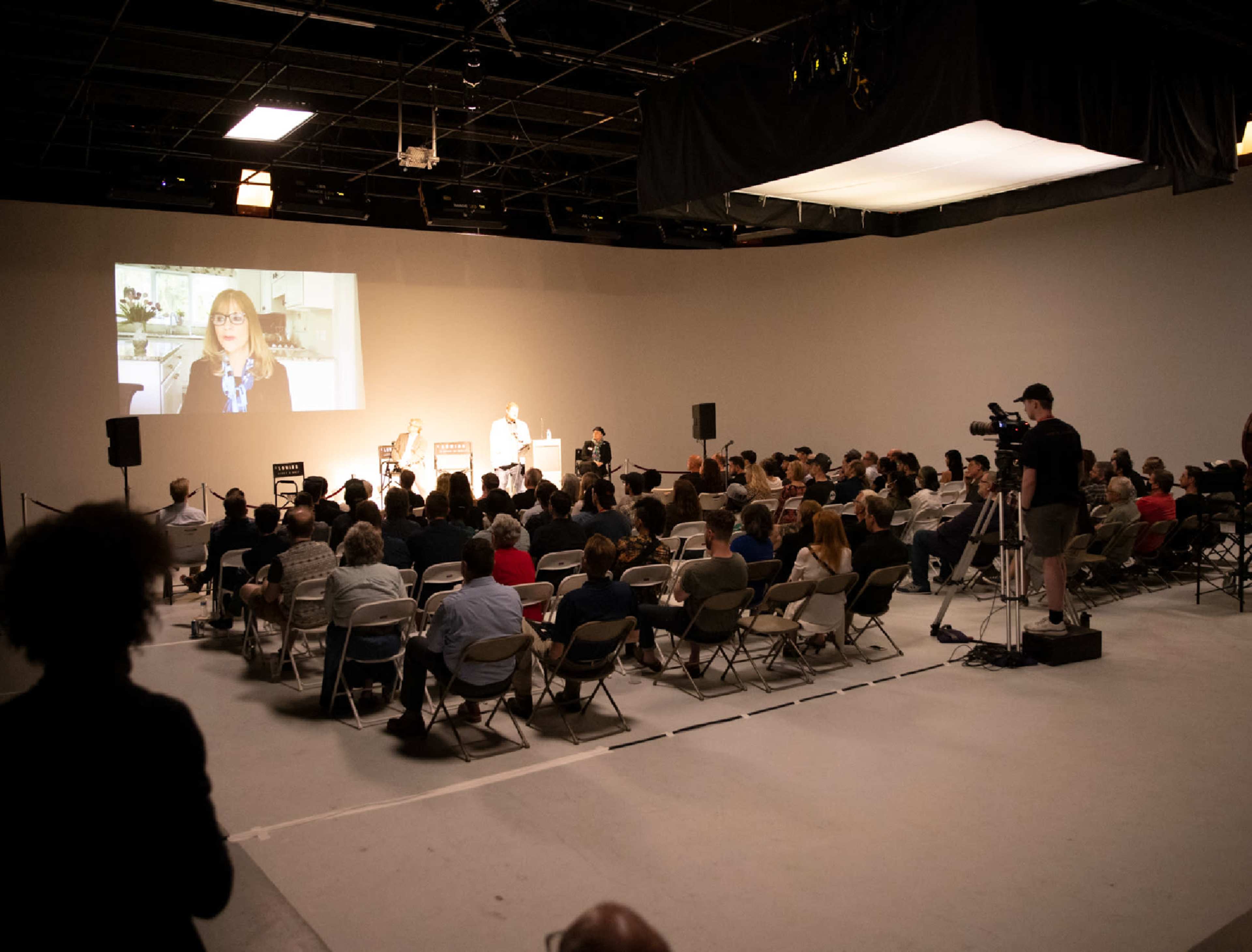 A large audience sits in a studio space with a presentation displayed on a screen at the front.