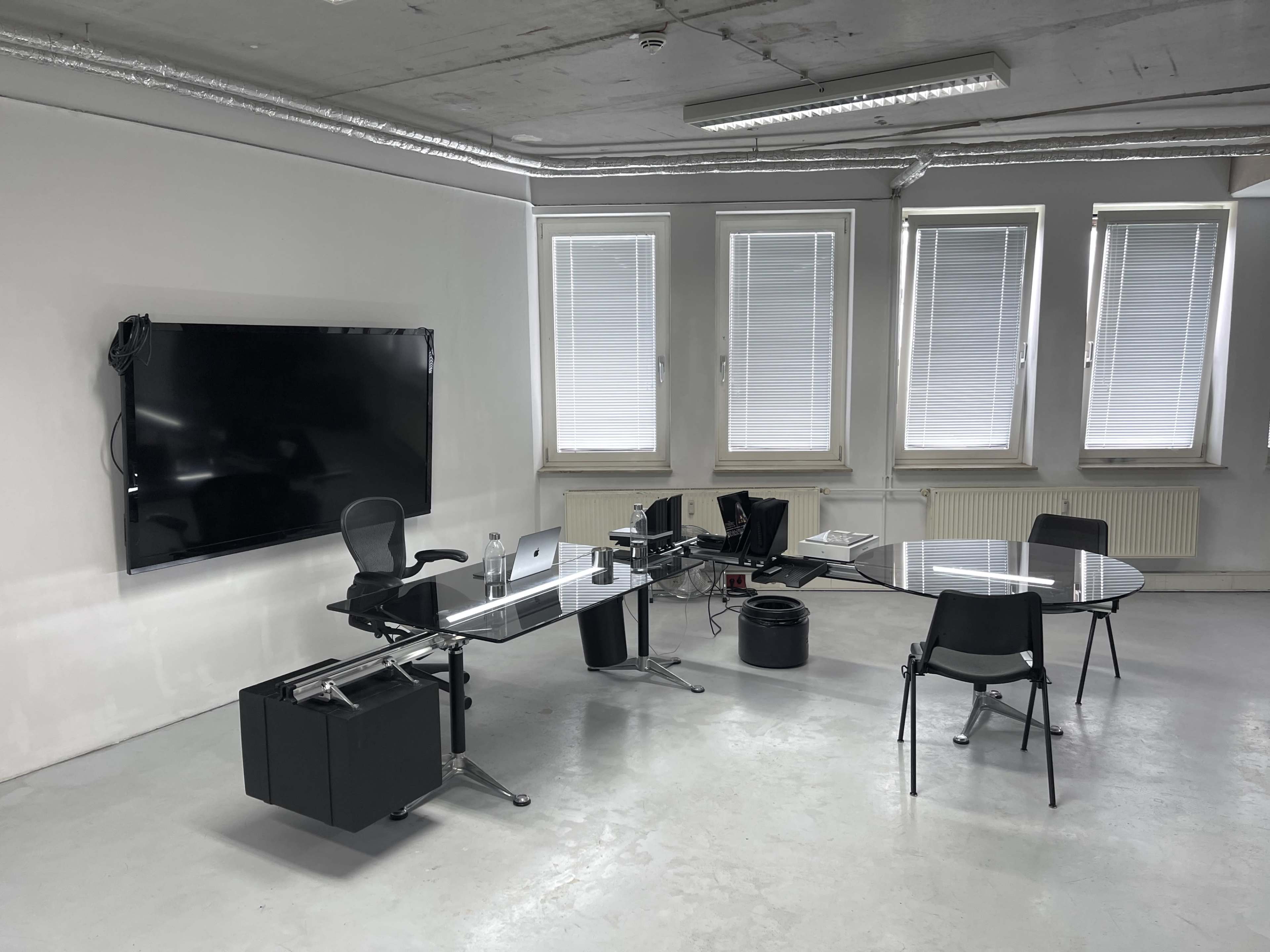 The image shows a minimalist office space featuring a glass desk with a chair, a round table with chairs, a large wall-mounted television, and multiple windows.