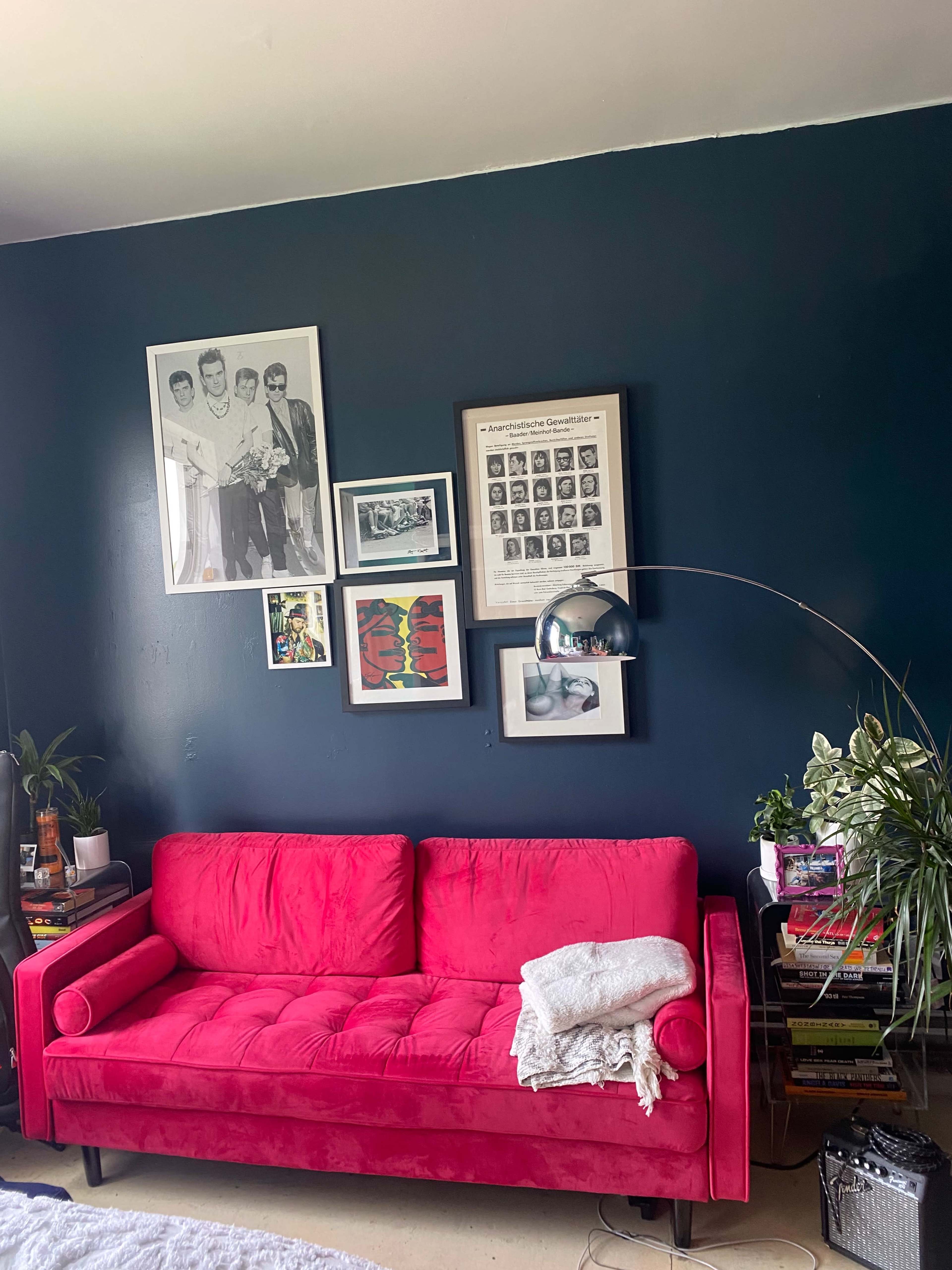 A bright pink sofa is positioned against a dark blue wall featuring a collection of framed artwork and photographs, with plants and books on a nearby table.