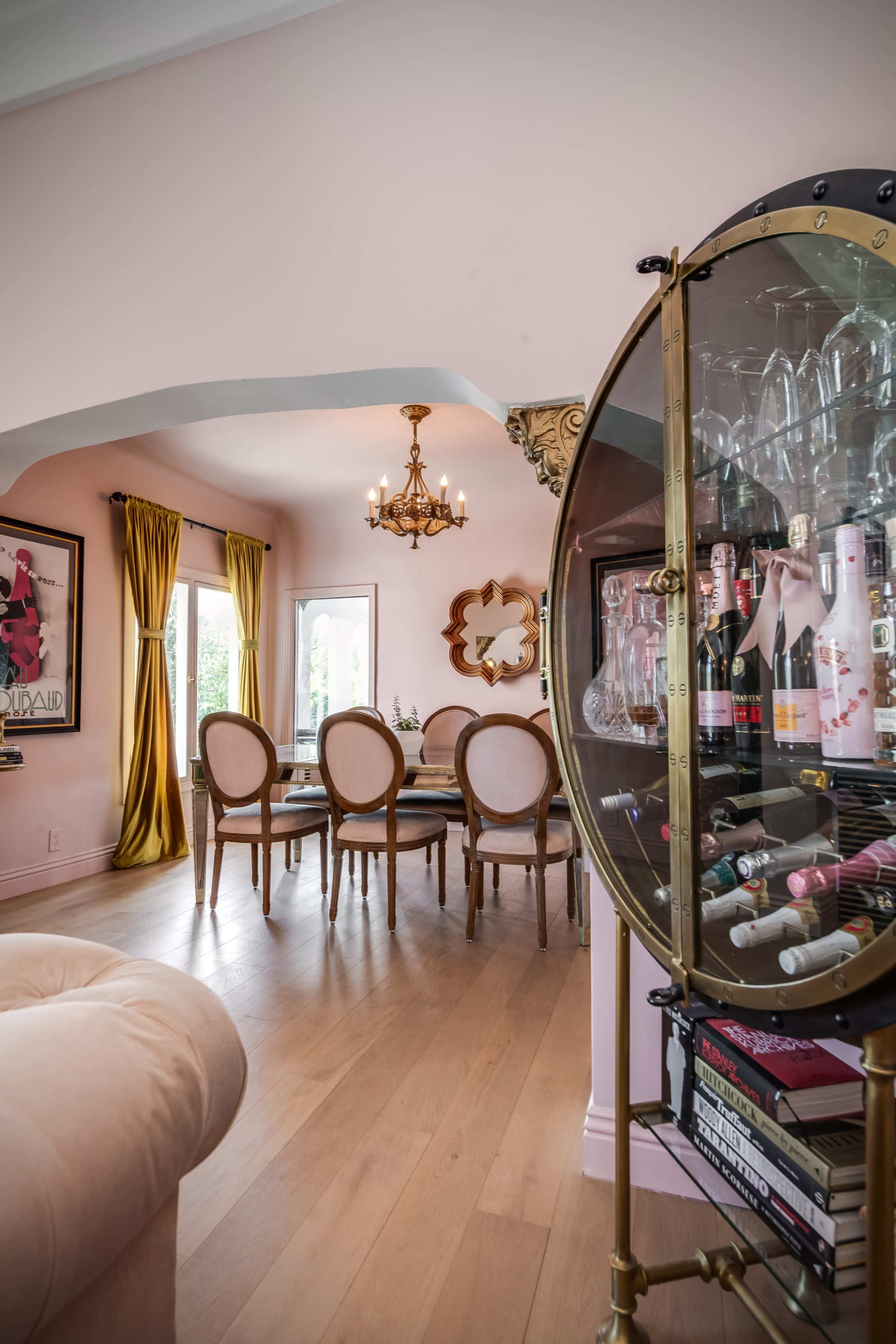 A dining area with a round table surrounded by eight chairs is visible, complemented by a bar cart displaying various bottles and glassware, with pink walls and decorative curtains.