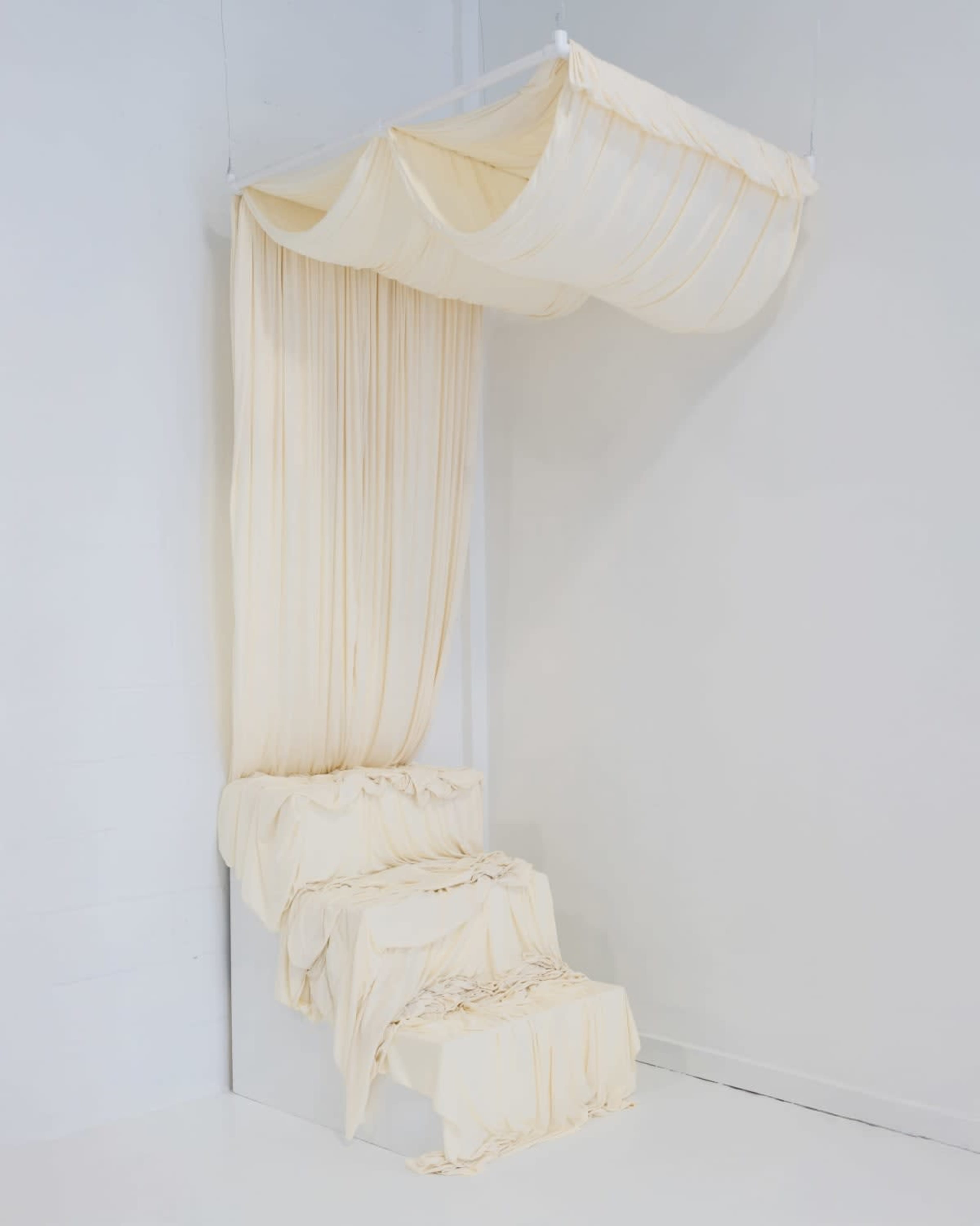 The image shows a minimalist installation featuring a white platform with cascading cream-colored fabric draped over it, resembling a flowing curtain.