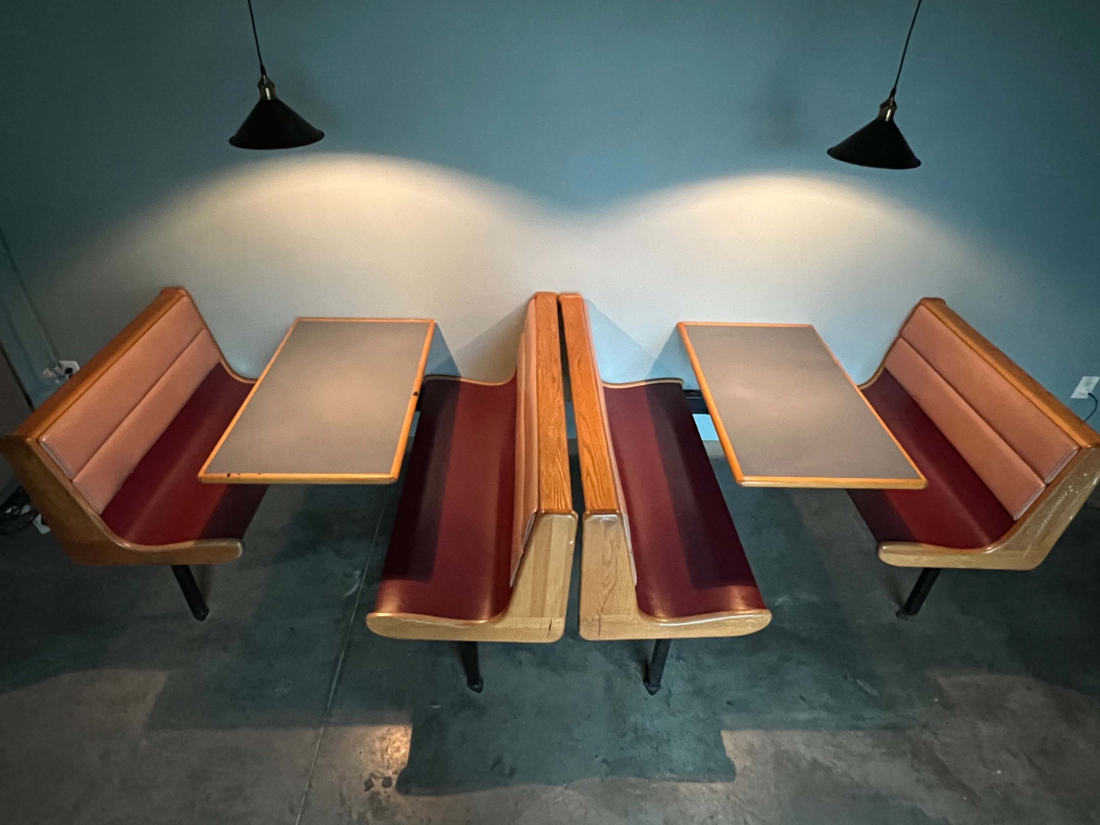 The image shows two wooden booth seats with maroon upholstery and two adjacent tables set in a minimalist dining area illuminated by pendant lights.