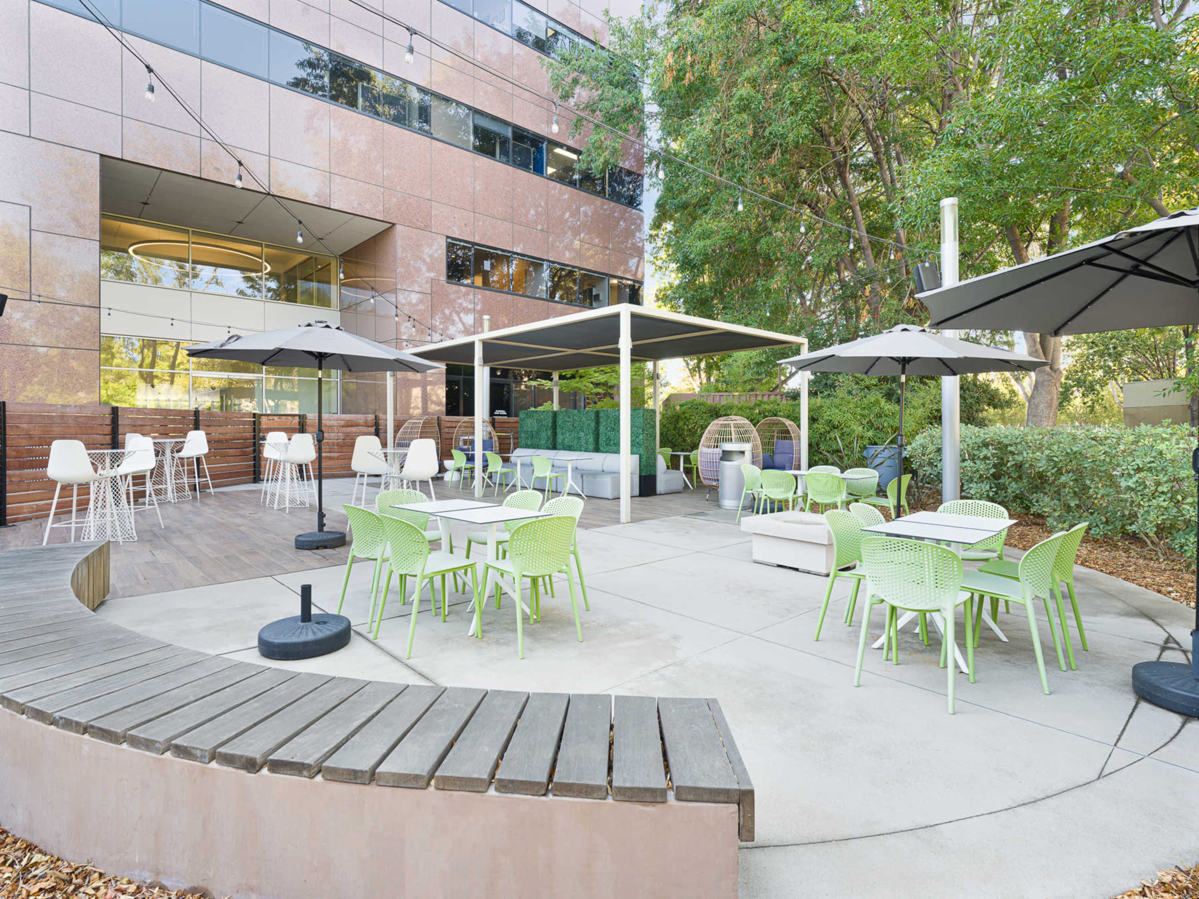 The image shows an outdoor seating area with green and white furniture, sunshades, and modern tables outside a building.