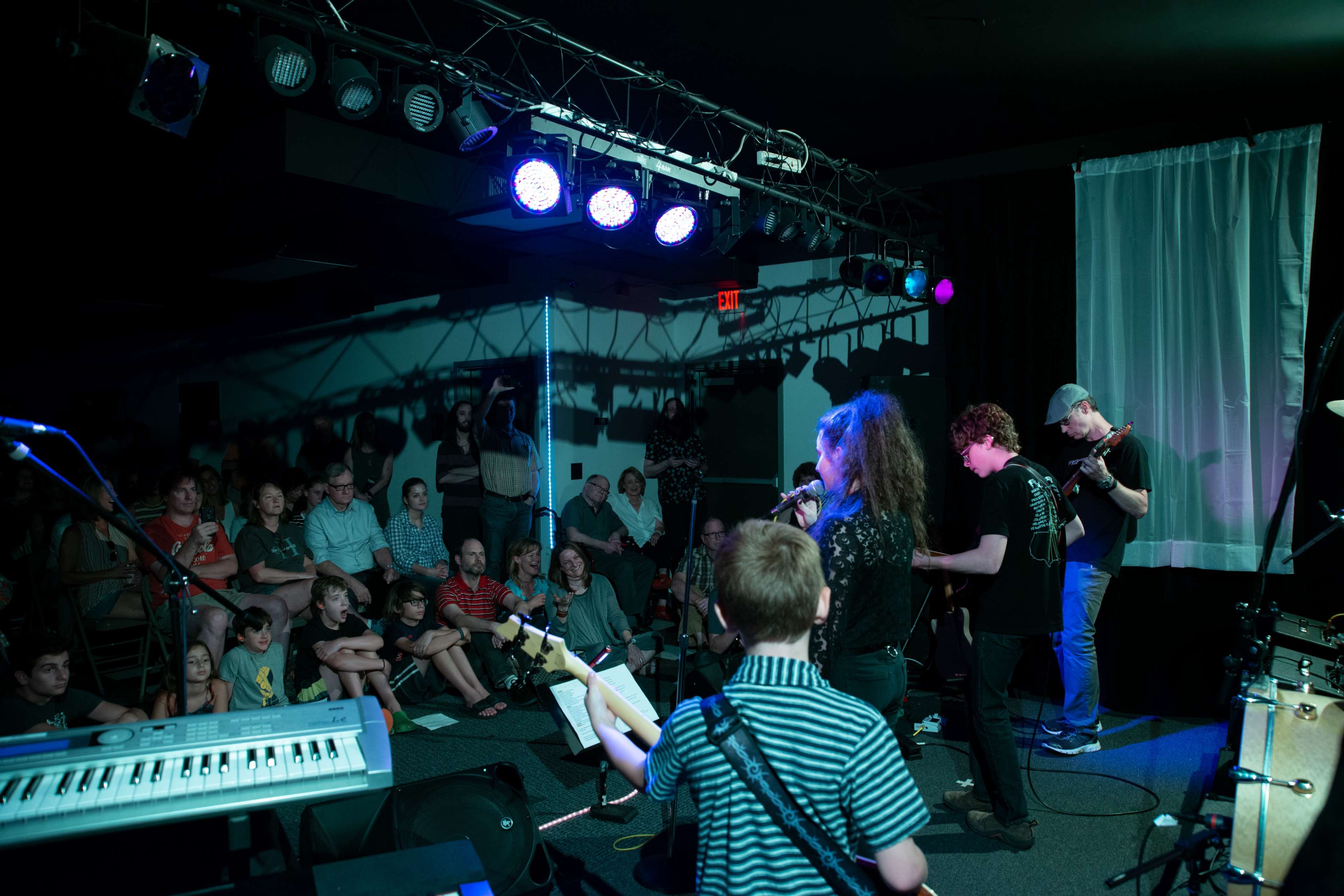 A group of young musicians performs on stage in front of an audience at a small venue.
