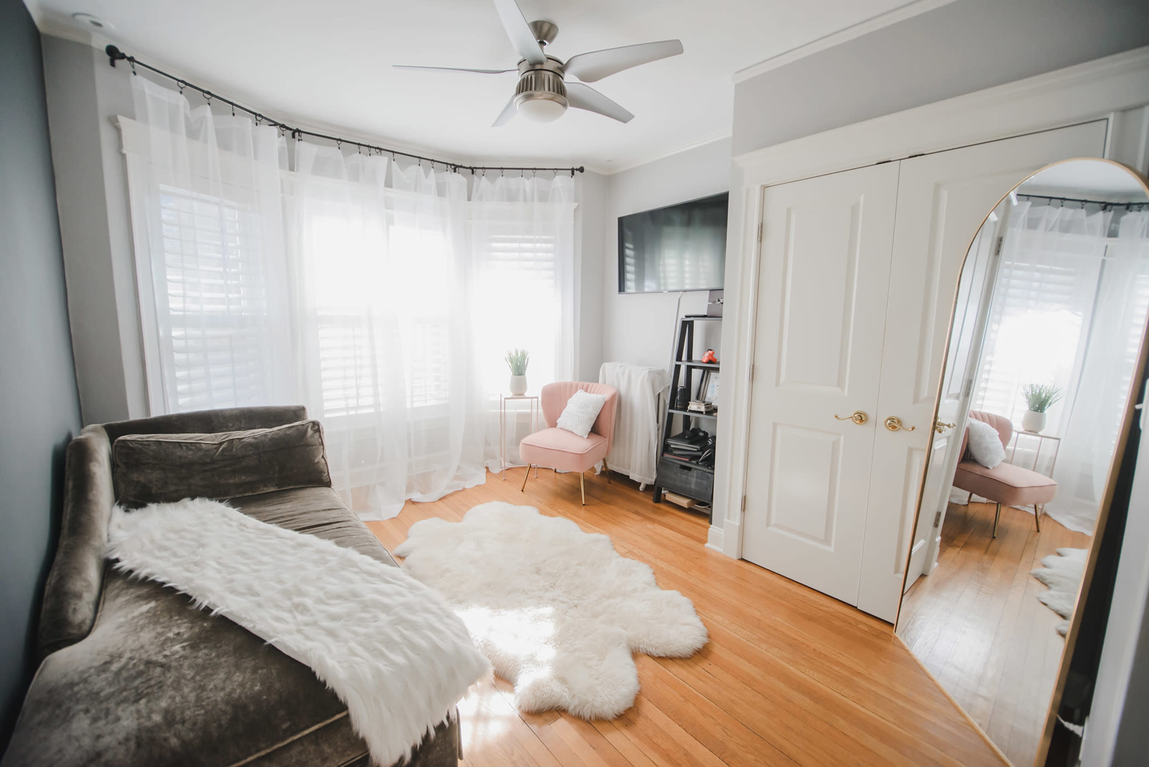 A well-lit room featuring a plush sofa, a light-colored rug, a small pink chair, and large windows dressed with sheer curtains.