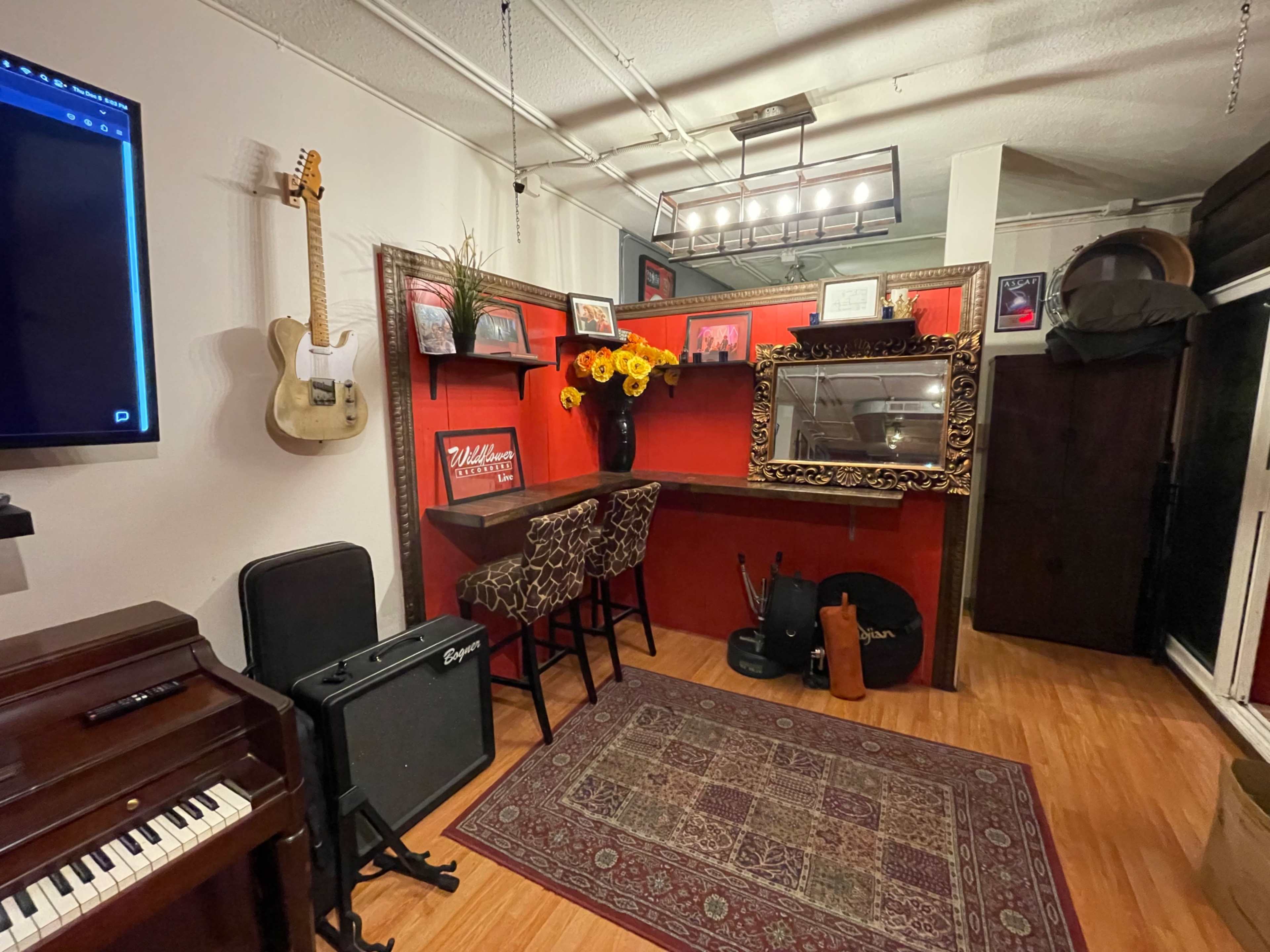 The image shows a cozy room featuring a red accent wall with a mirror, a guitar hanging, a small bar area with stools, and various musical instruments.