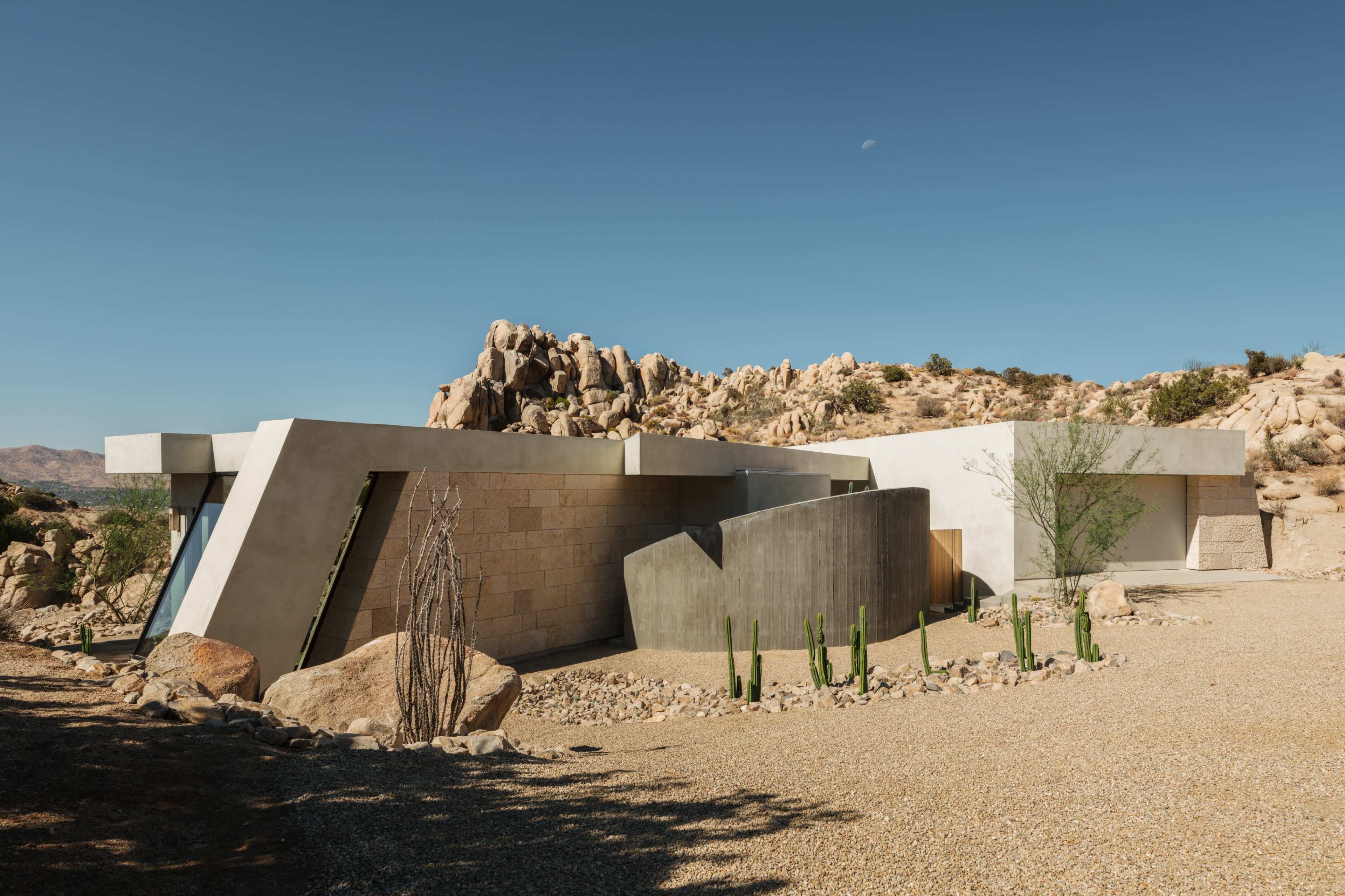 The image features a modern architectural structure with angular lines, situated among rocky terrain and sparse vegetation in a desert landscape.