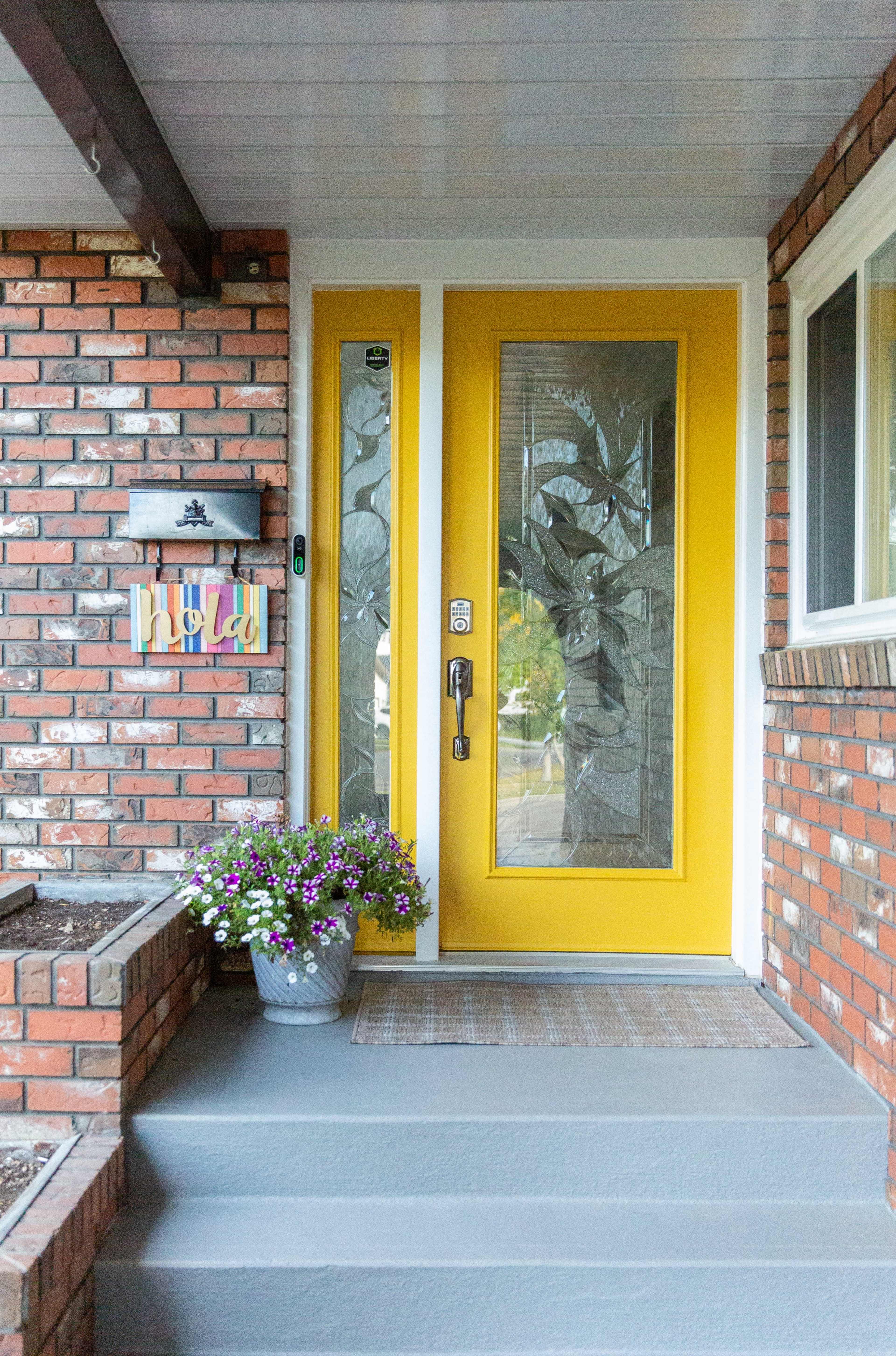 The image shows a bright yellow front door decorated with a patterned glass insert, flanked by potted flowers and a welcome sign on a brick home.